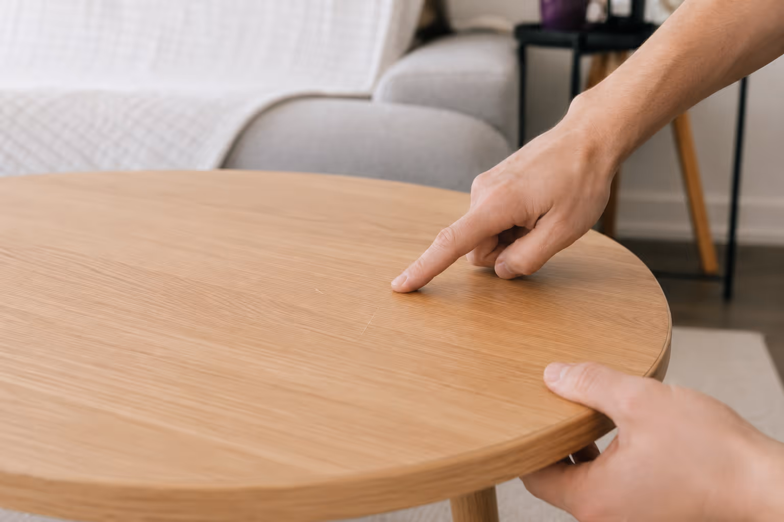 Person pointing at a small scratch on a round wooden table in a living room.