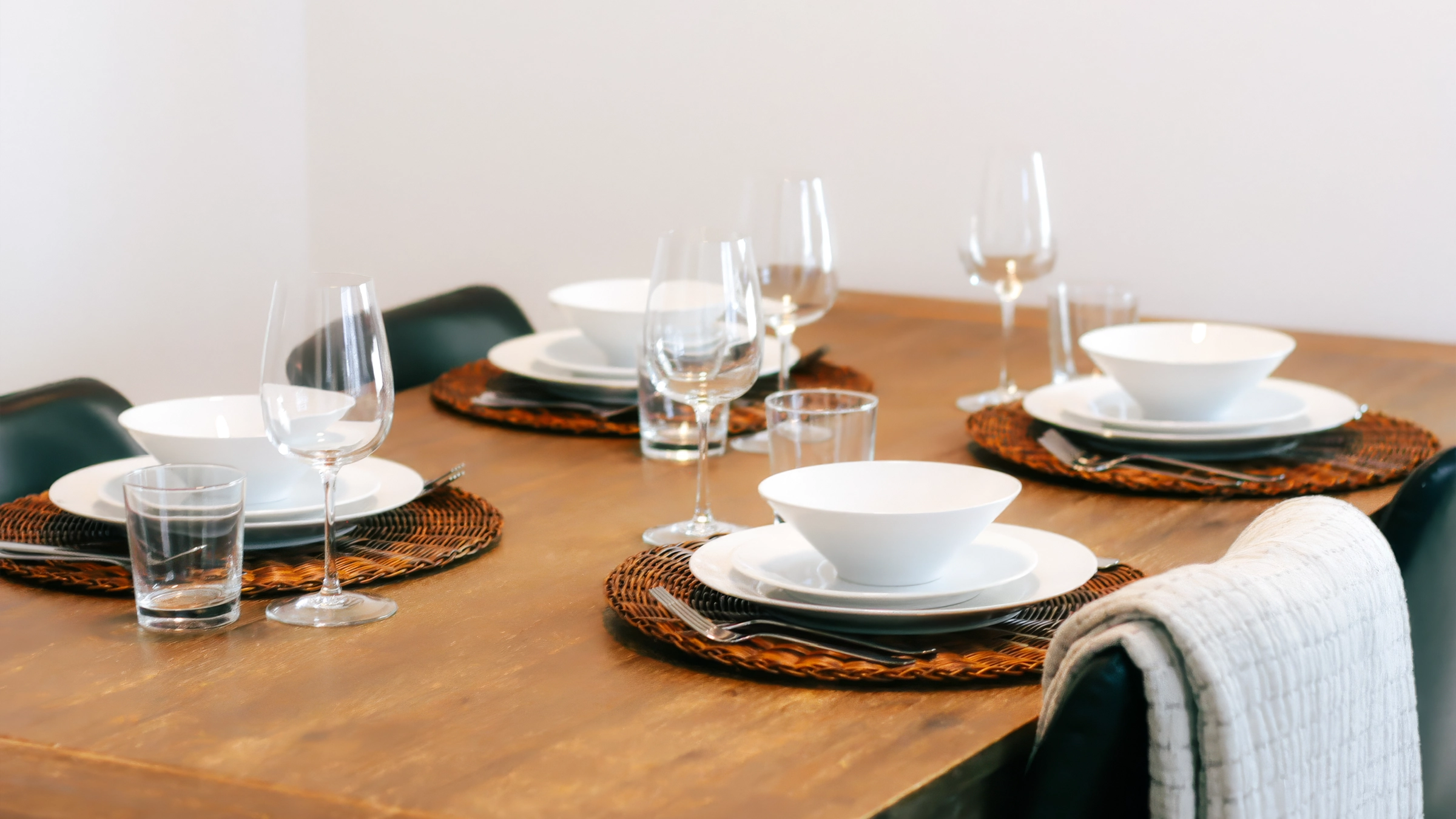Wooden dining table set with white bowls, plates, glasses, and woven placemats for four people.