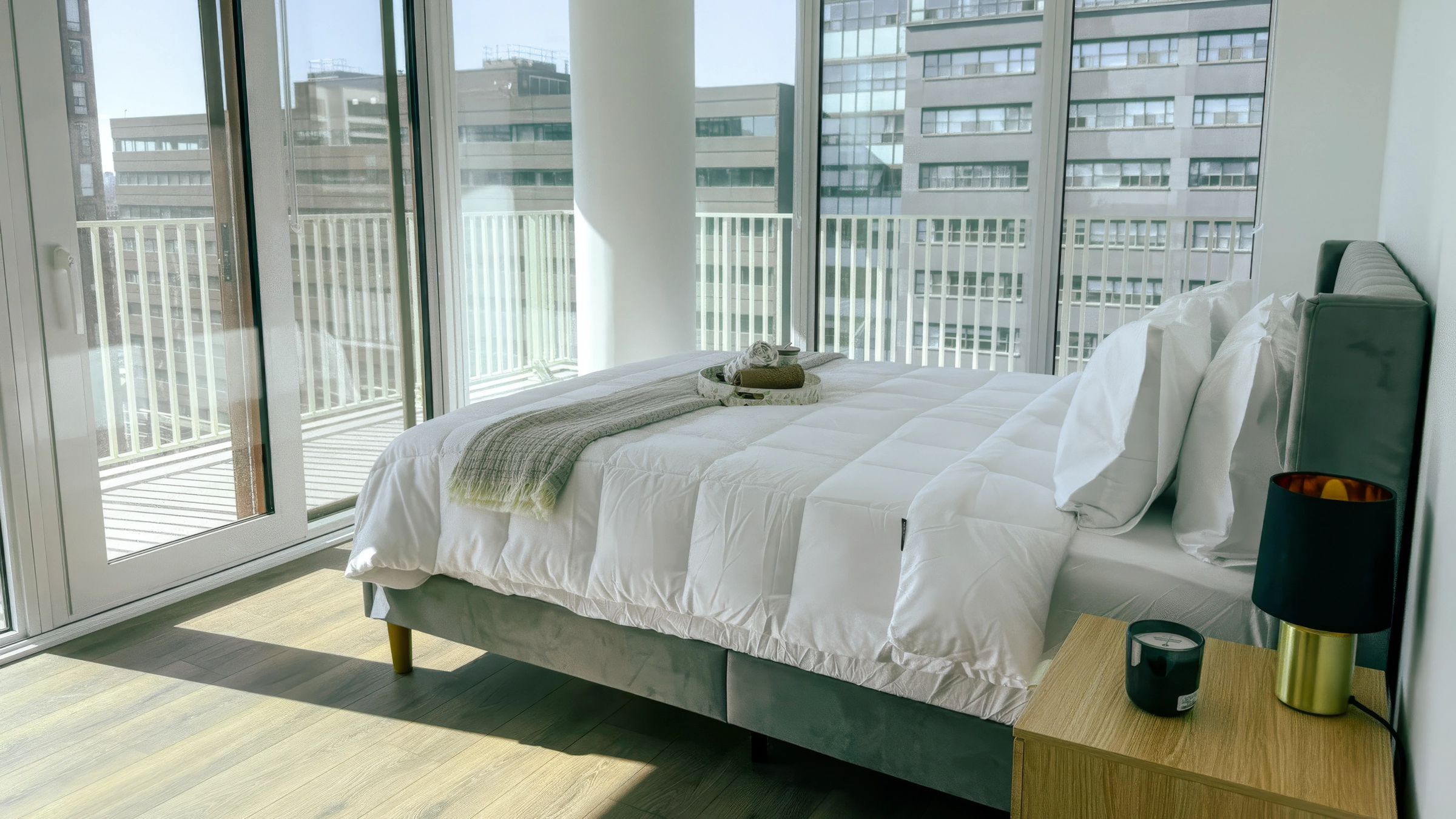 Modern bedroom with a made bed, gray headboard, wooden nightstand, and large windows with city view.
