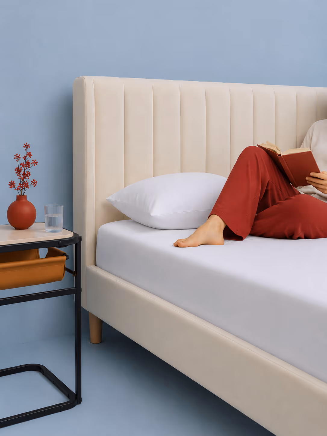 Person in red pants reading on a beige bed with white pillow next to a table with vase and water glass.