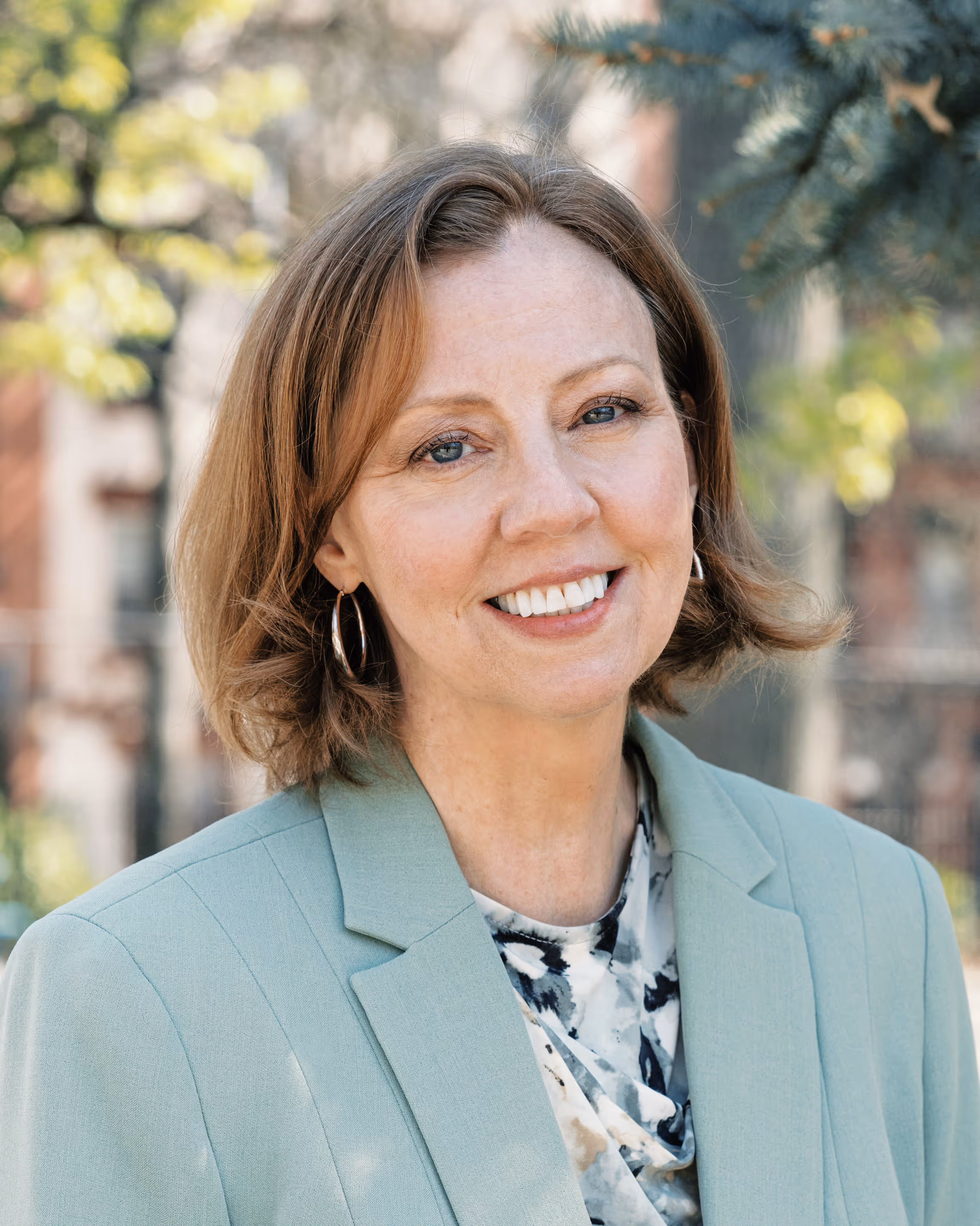 Smiling middle-aged woman with short light brown hair wearing a light green blazer and patterned blouse standing outdoors.