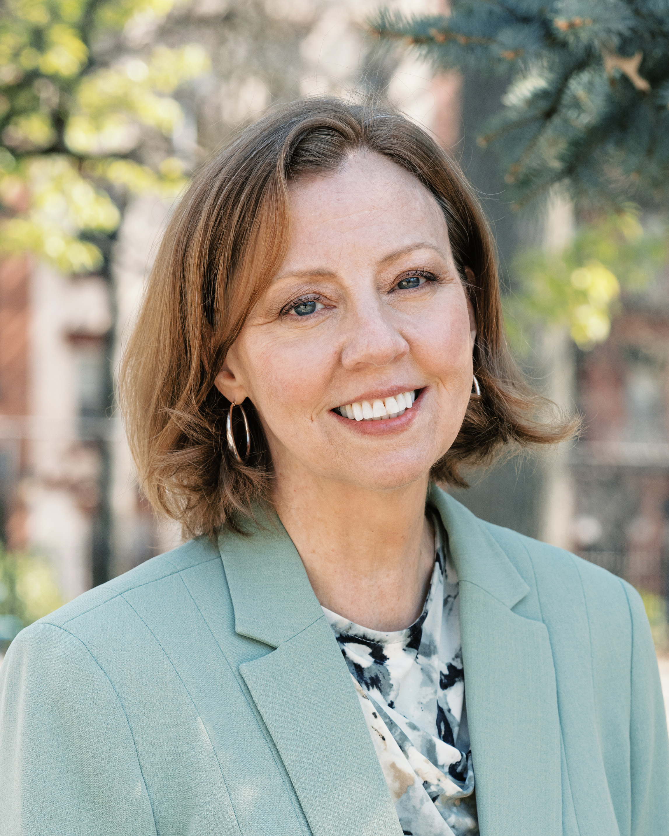 Smiling middle-aged woman with short light brown hair wearing a light green blazer and patterned blouse standing outdoors.