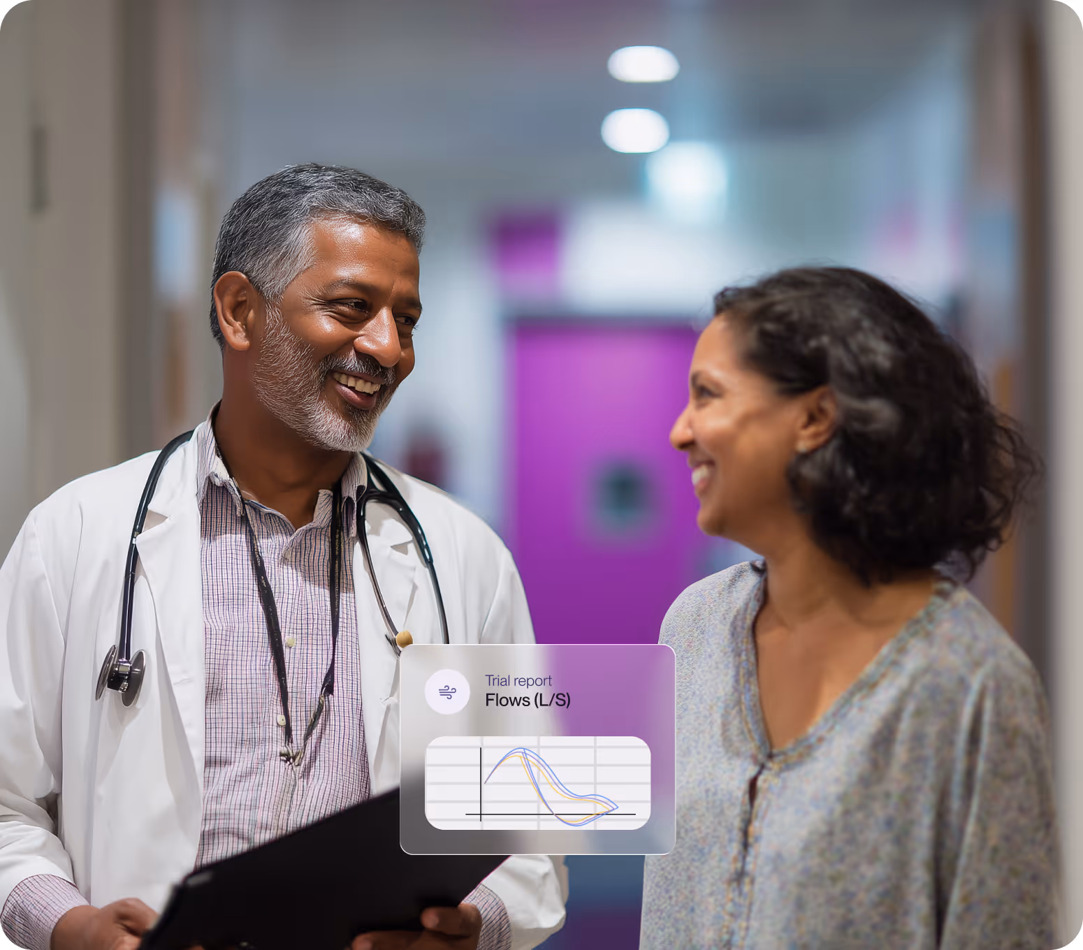 Smiling male doctor with a stethoscope talks to a female patient in a hallway.