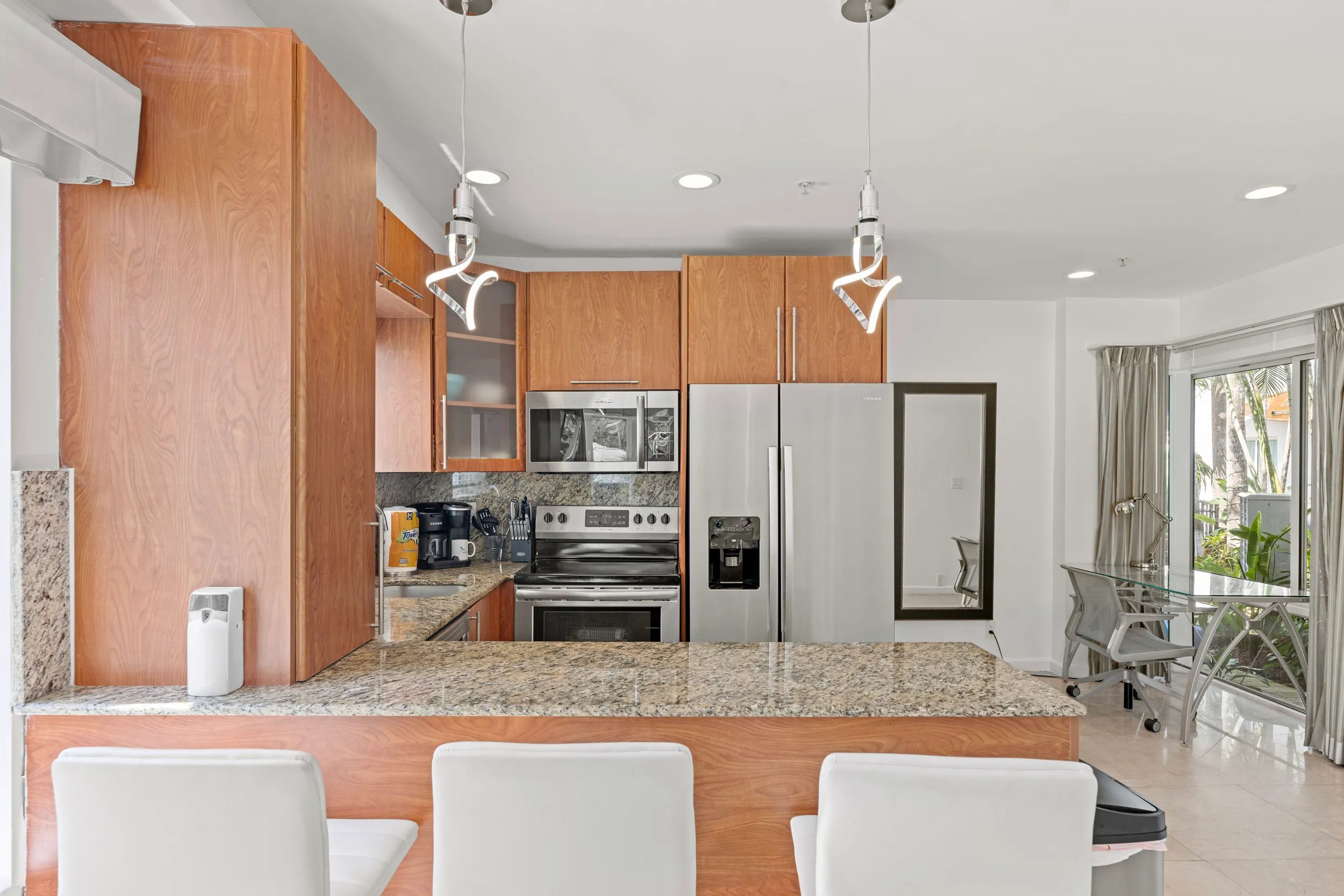 Modern kitchen with wooden cabinets, granite countertops, stainless steel refrigerator, oven, and microwave, three white bar stools at the counter, and an adjacent glass dining table with chairs near sliding glass doors.