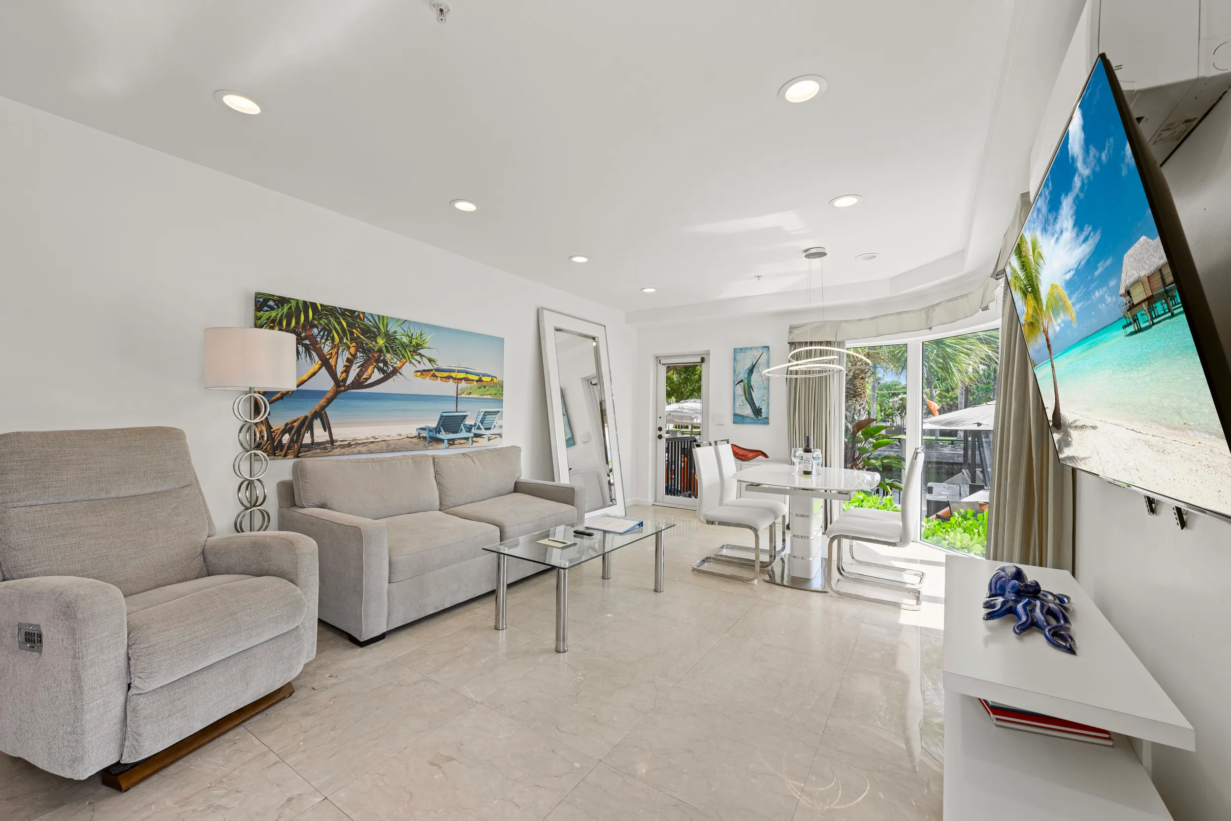 Modern living room with beige sofa, recliner, glass coffee table, dining area with white chairs, large mirror, and tropical beach paintings.
