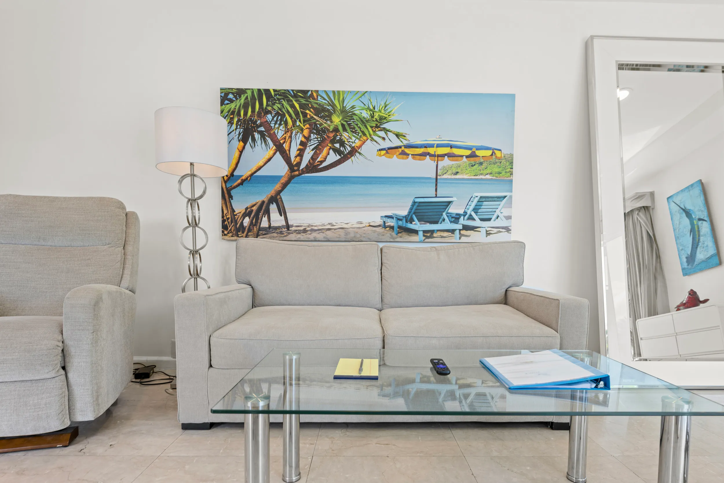 Modern living room with beige couch, glass coffee table, a scenic beach wall painting, and a large floor mirror.