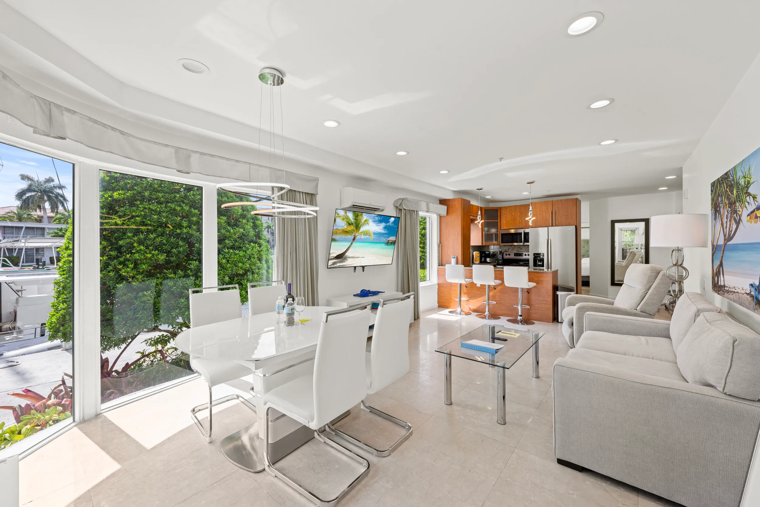 Bright open living and dining area with white furniture, glass coffee table, modern kitchen with wooden cabinets, and large windows showing a green outdoor view.