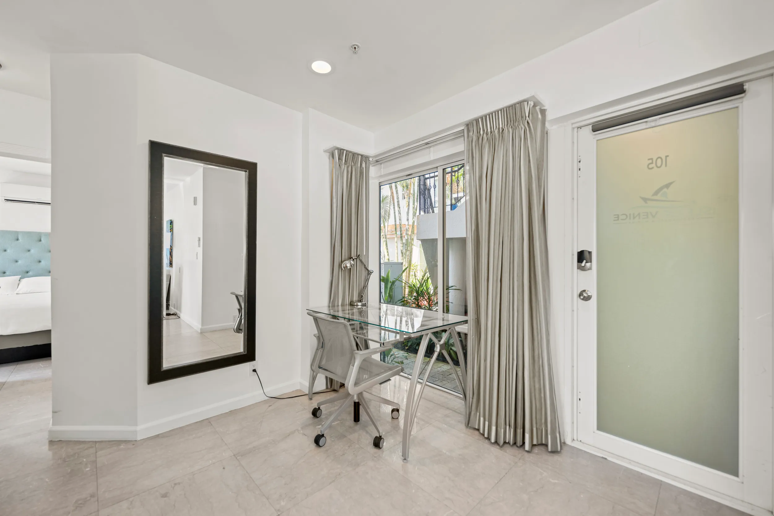 Modern bright office corner with glass desk, gray chair on casters, wall mirror, and large window with curtains.