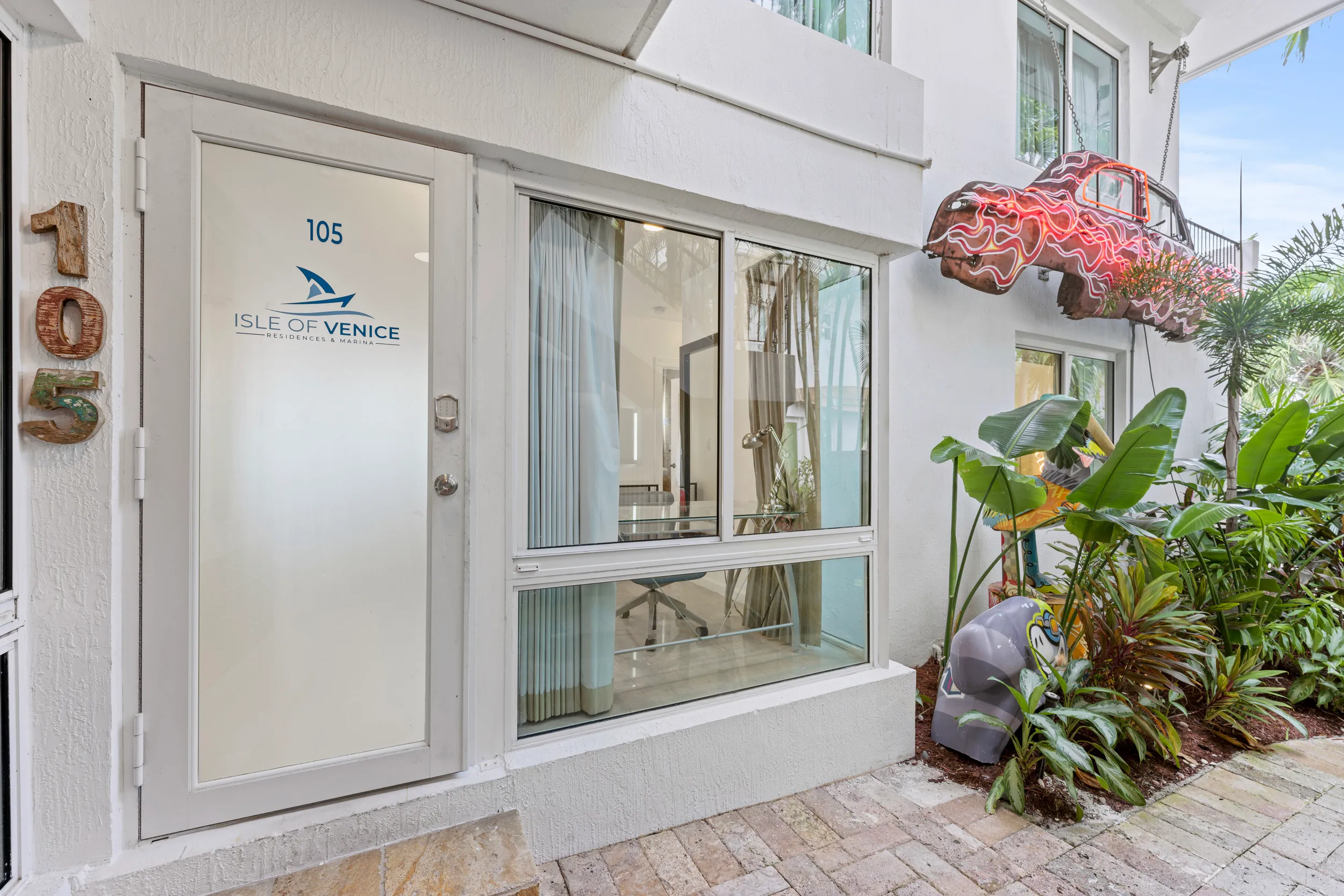 Entrance door numbered 105 with Isle of Venice Residences & Marina logo next to a window and hanging car sculpture with neon light accents.
