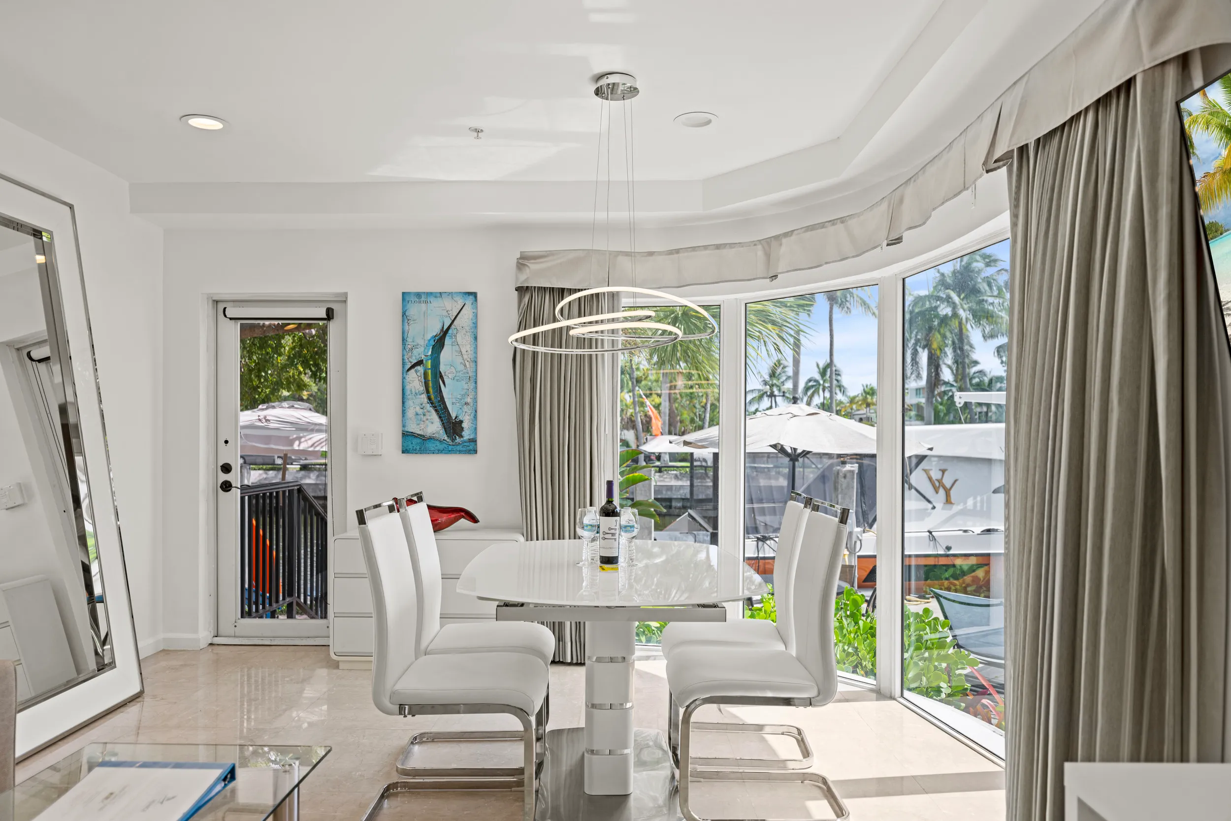 Modern dining area with white chairs and table by large windows showing palm trees and boats outside.
