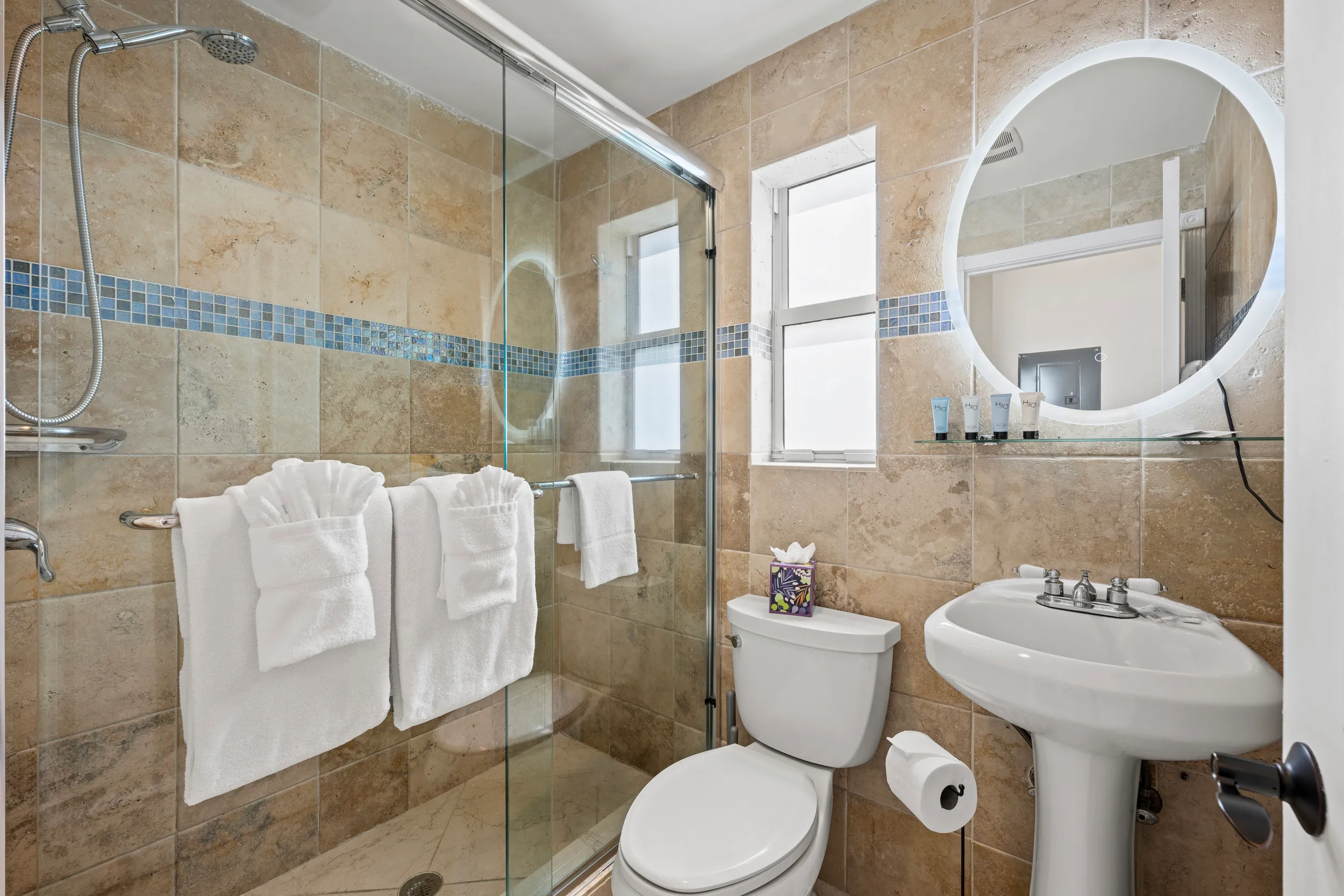 Small bathroom with beige tiled walls, glass shower with two white towels, white toilet with tissue box, and pedestal sink with round backlit mirror.