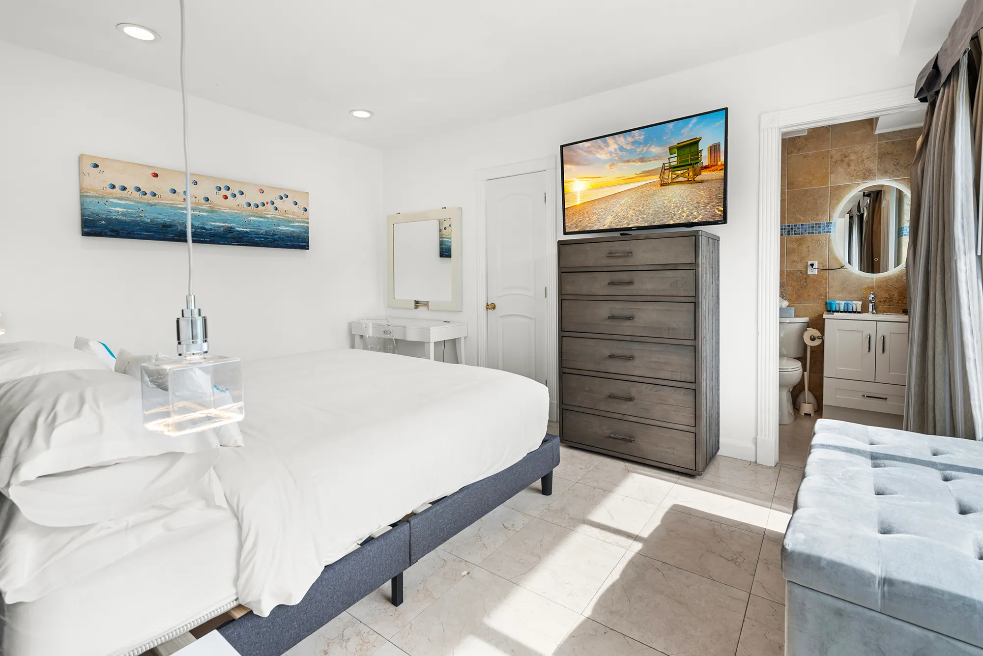 Bright bedroom with white bedding, a wooden dresser topped with a TV, a vanity mirror, and an open door leading to a bathroom.