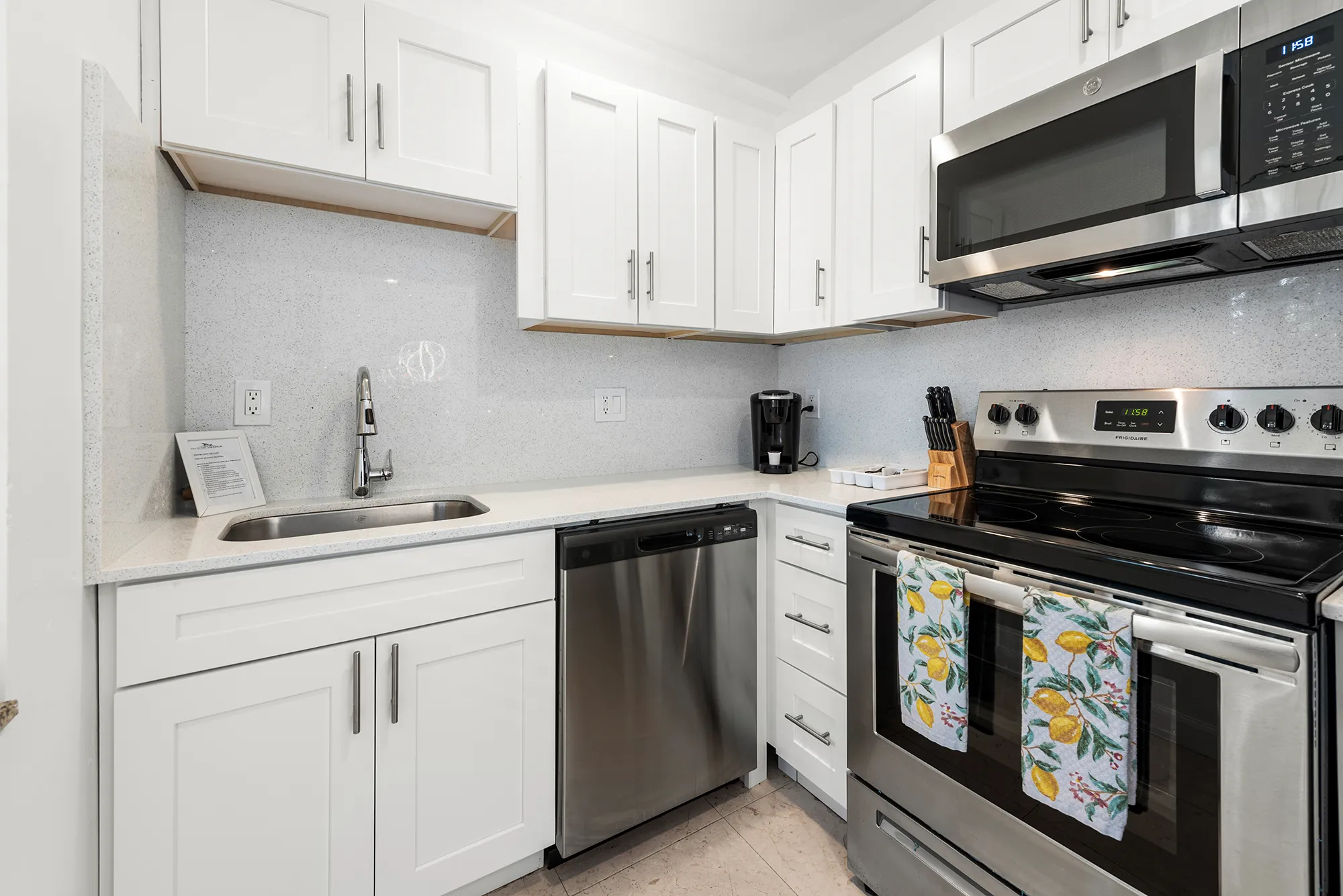 Modern white kitchen corner with stainless steel dishwasher, stove with lemon-patterned towels, microwave, sink, and coffee maker.