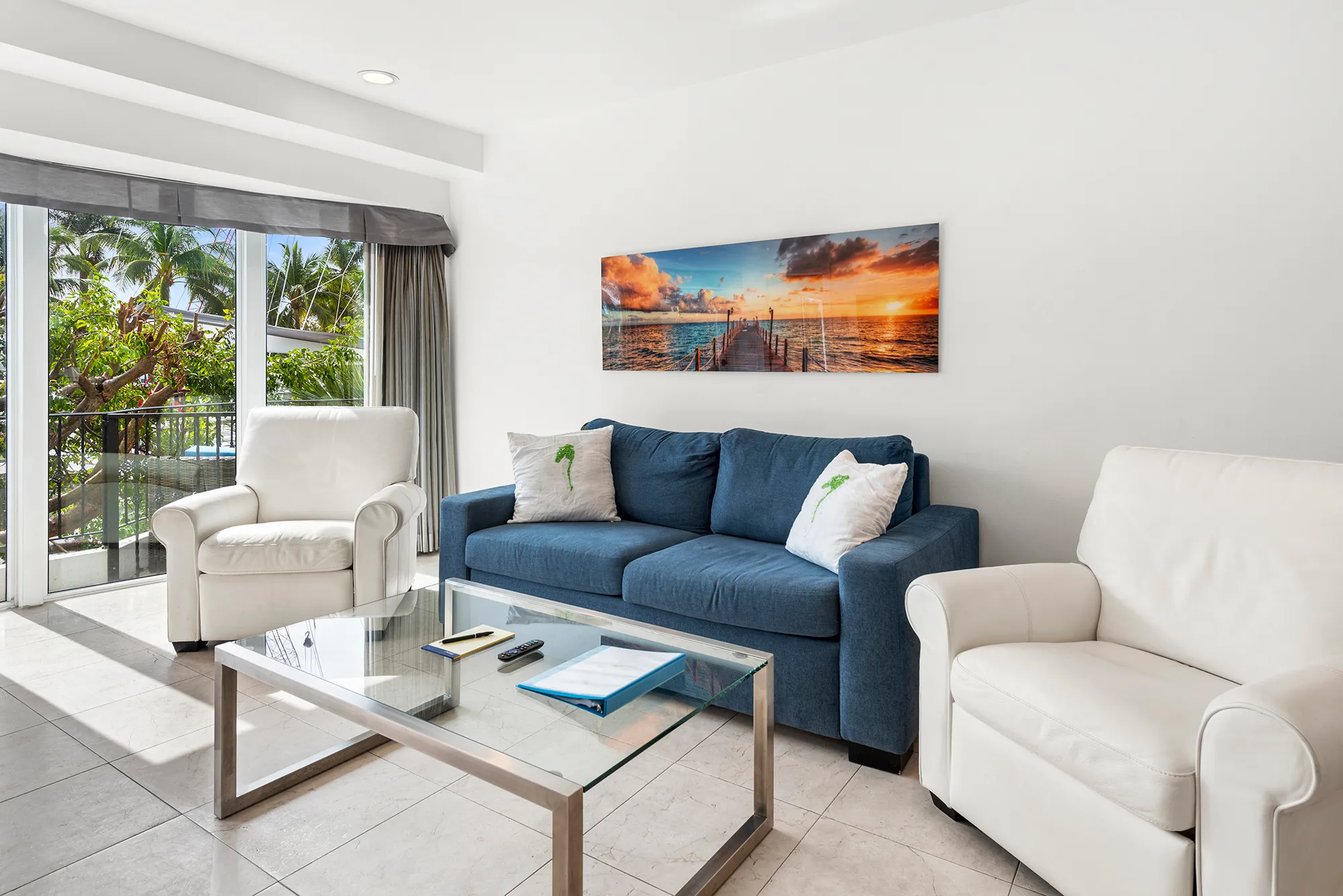 Bright living room with a blue sofa, two white armchairs, a glass coffee table, and a sunset pier wall art above the sofa.
