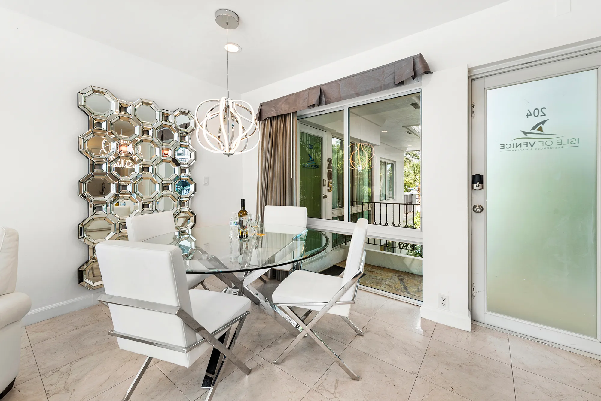 Modern dining area with glass round table, four white cushioned chairs with metal legs, a decorative geometric mirror on the wall, and a pendant light fixture, next to a frosted glass door and large window.