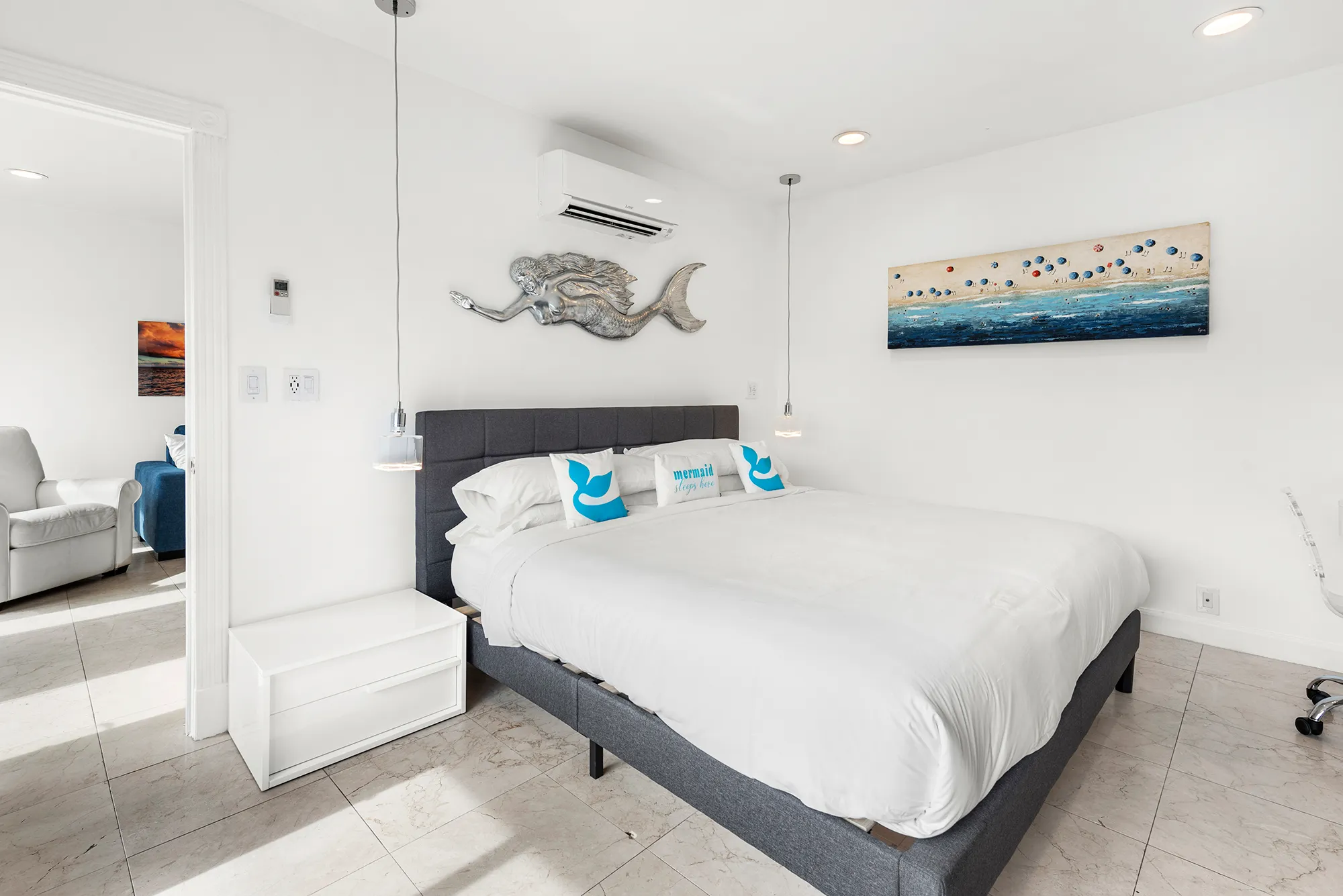 Bright bedroom with a large bed featuring white bedding and three decorative pillows, a silver mermaid wall sculpture above the headboard, and a coastal painting on the adjacent wall.