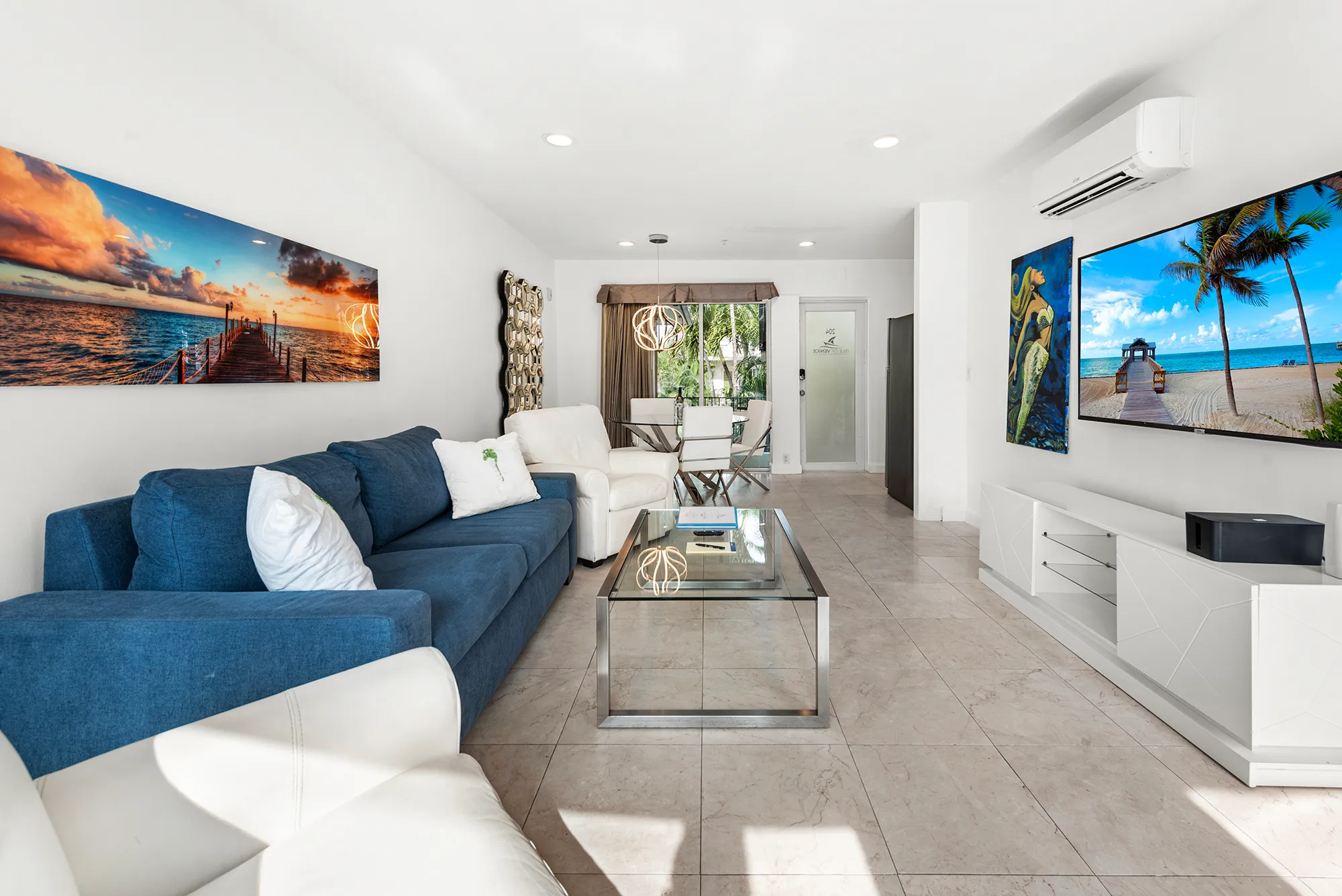 Modern living room with blue and white sofas, glass coffee table, wall-mounted TV showing beach scene, and dining area with glass table and white chairs.