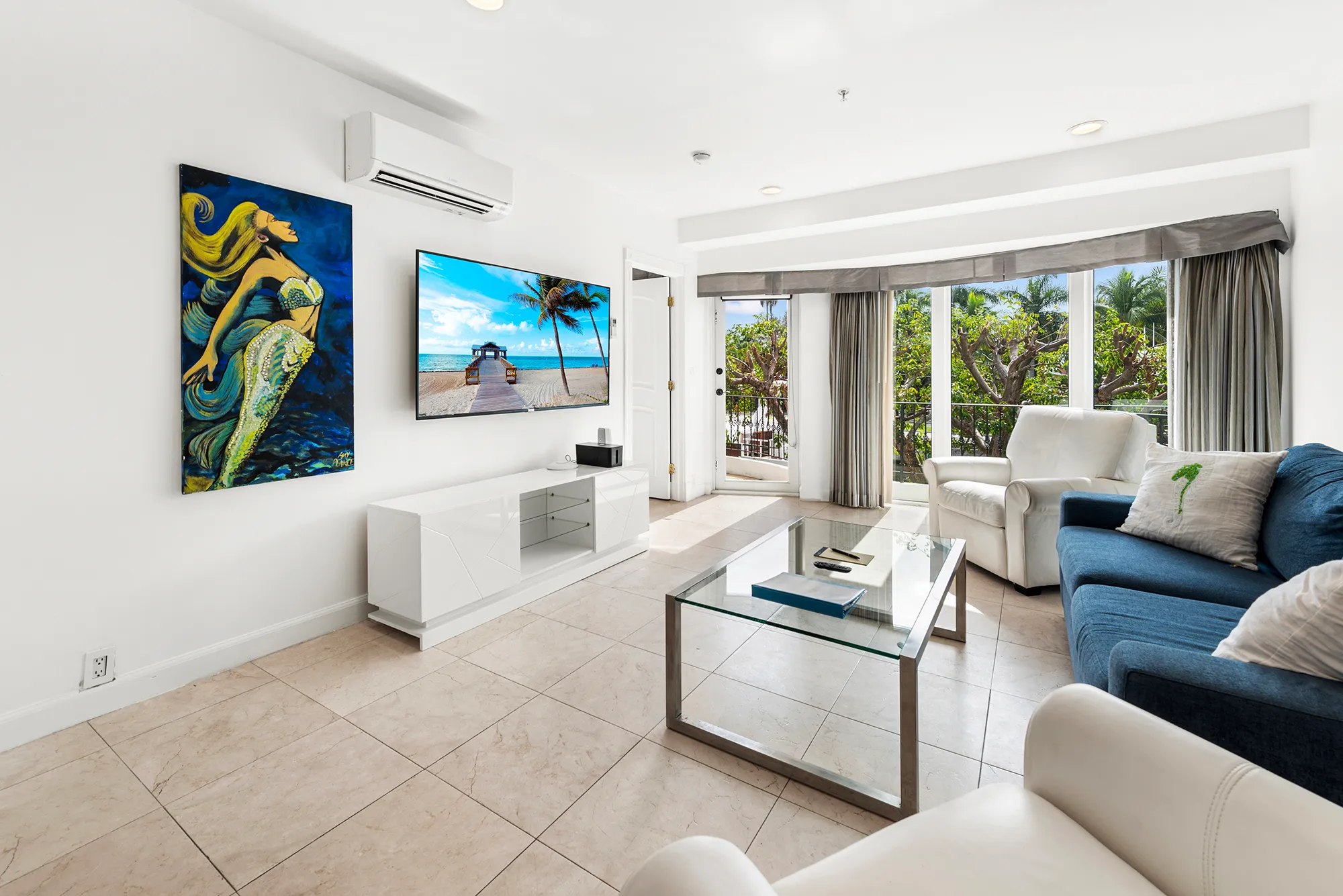Bright living room with white walls, a blue sofa, white armchairs, glass coffee table, wall-mounted TV showing a beach scene, and a painting of a mermaid.