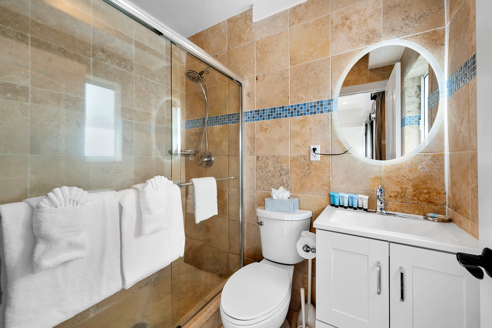 Clean bathroom with a glass shower enclosure, white towels on the door, a toilet with tissue box on top, and a white vanity with a lit round mirror.