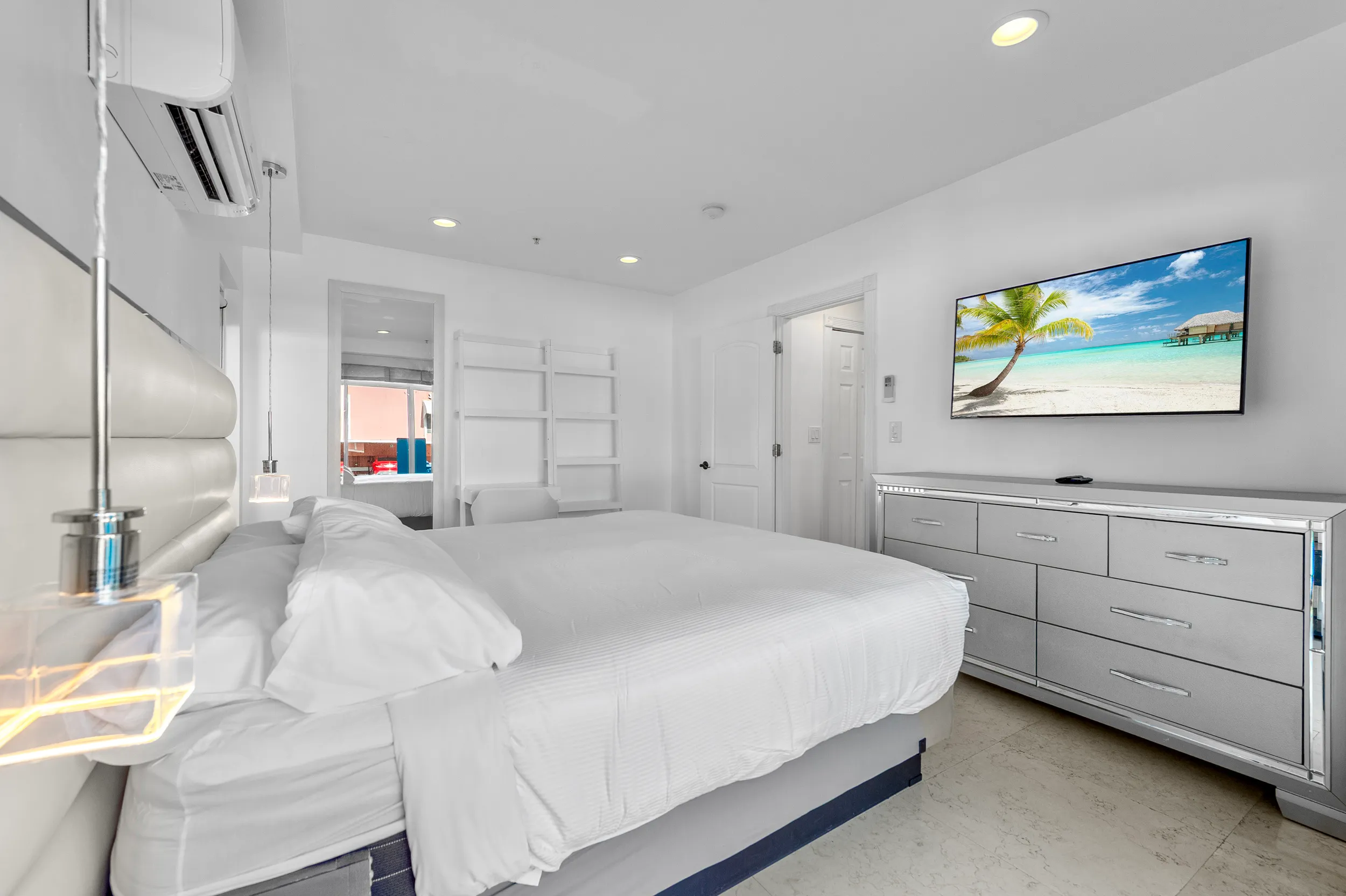 Bright bedroom with a large white bed, hanging lights, mirrored dresser, and a TV showing a tropical beach.