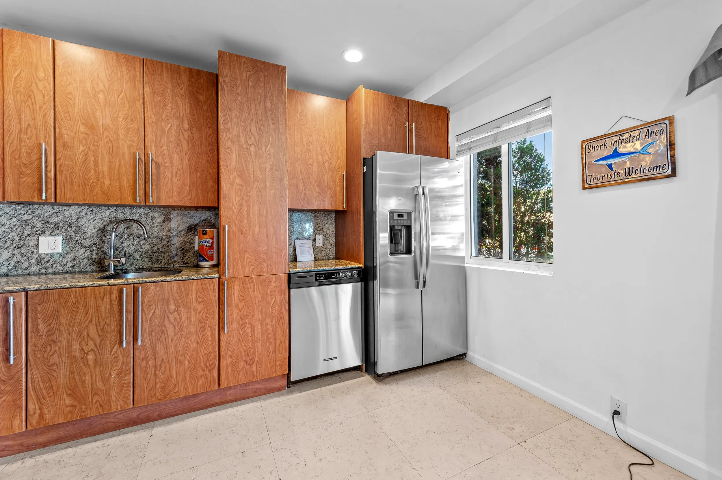 Modern kitchen with wooden cabinets, granite countertop, stainless steel refrigerator and dishwasher, and a window with a shark-themed sign on the wall.