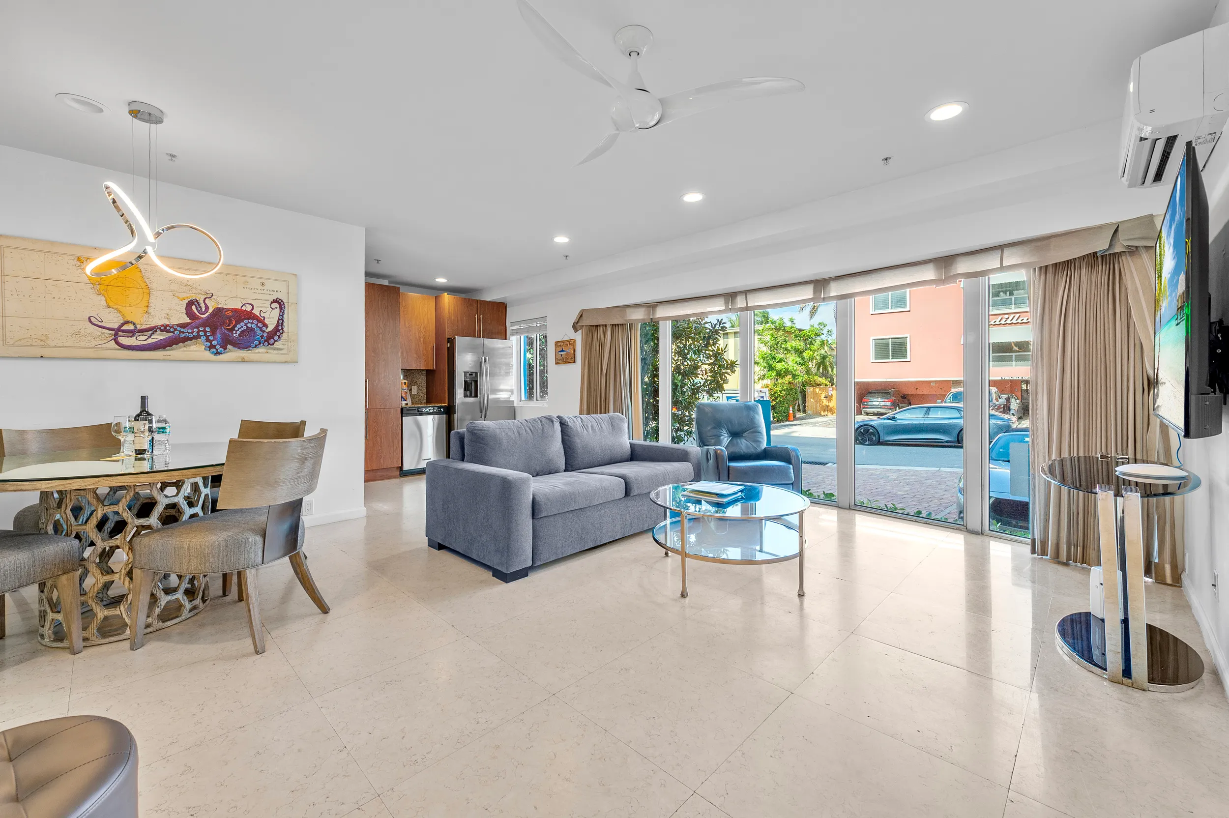 Bright living room with large sliding glass doors, a gray sofa and armchair, round glass coffee table, dining area with table and chairs, and modern ceiling fan.