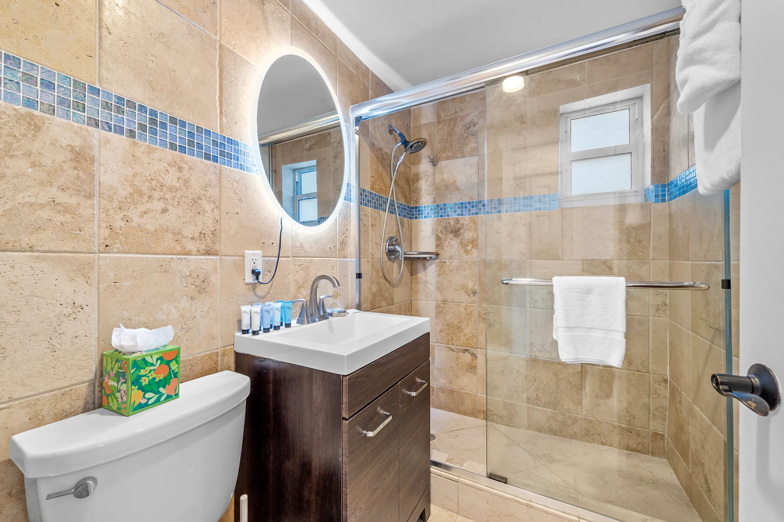 Modern bathroom with beige tiled walls, a glass-enclosed shower with blue tile border, a wooden vanity with a white sink, and a round backlit mirror.