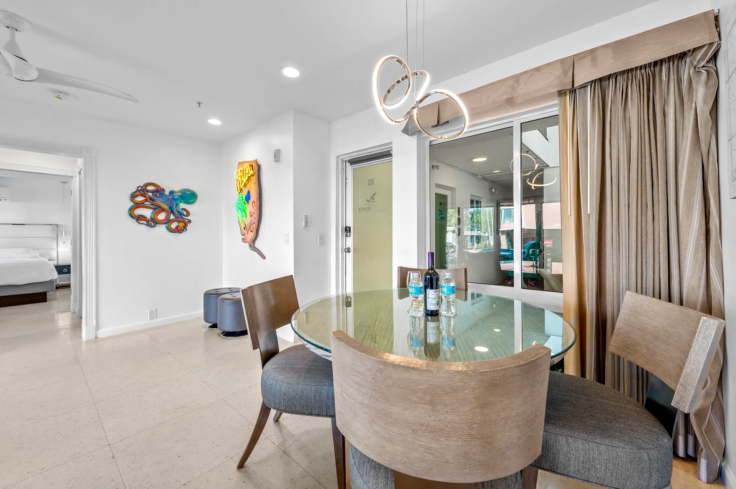 Modern dining area with round glass table, four cushioned chairs, water bottles, wine bottle, and unique wall art of an octopus and a neon face.