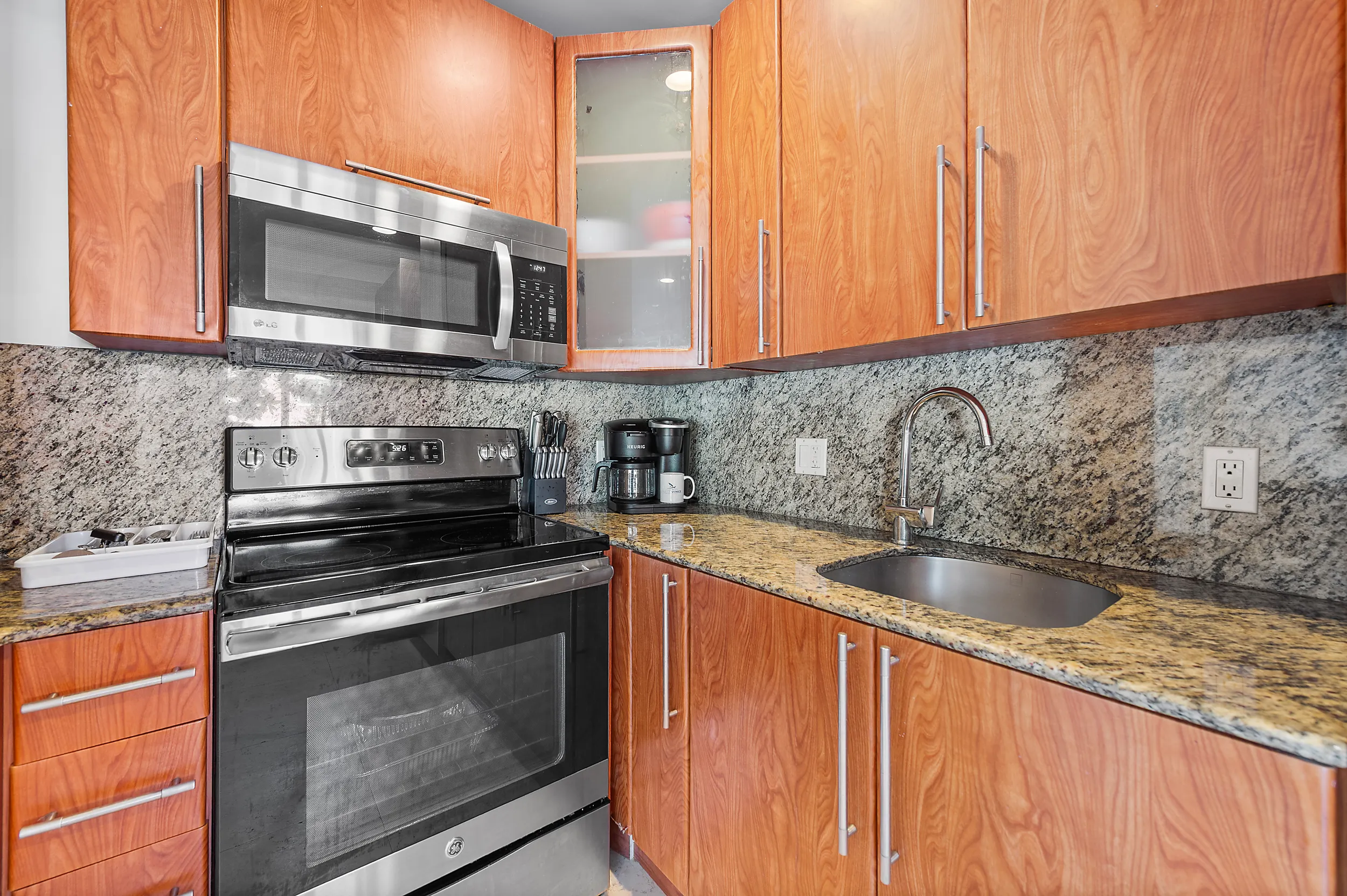 Modern kitchen corner with stainless steel electric stove, microwave, granite countertops, wooden cabinets, coffee maker, and sink.