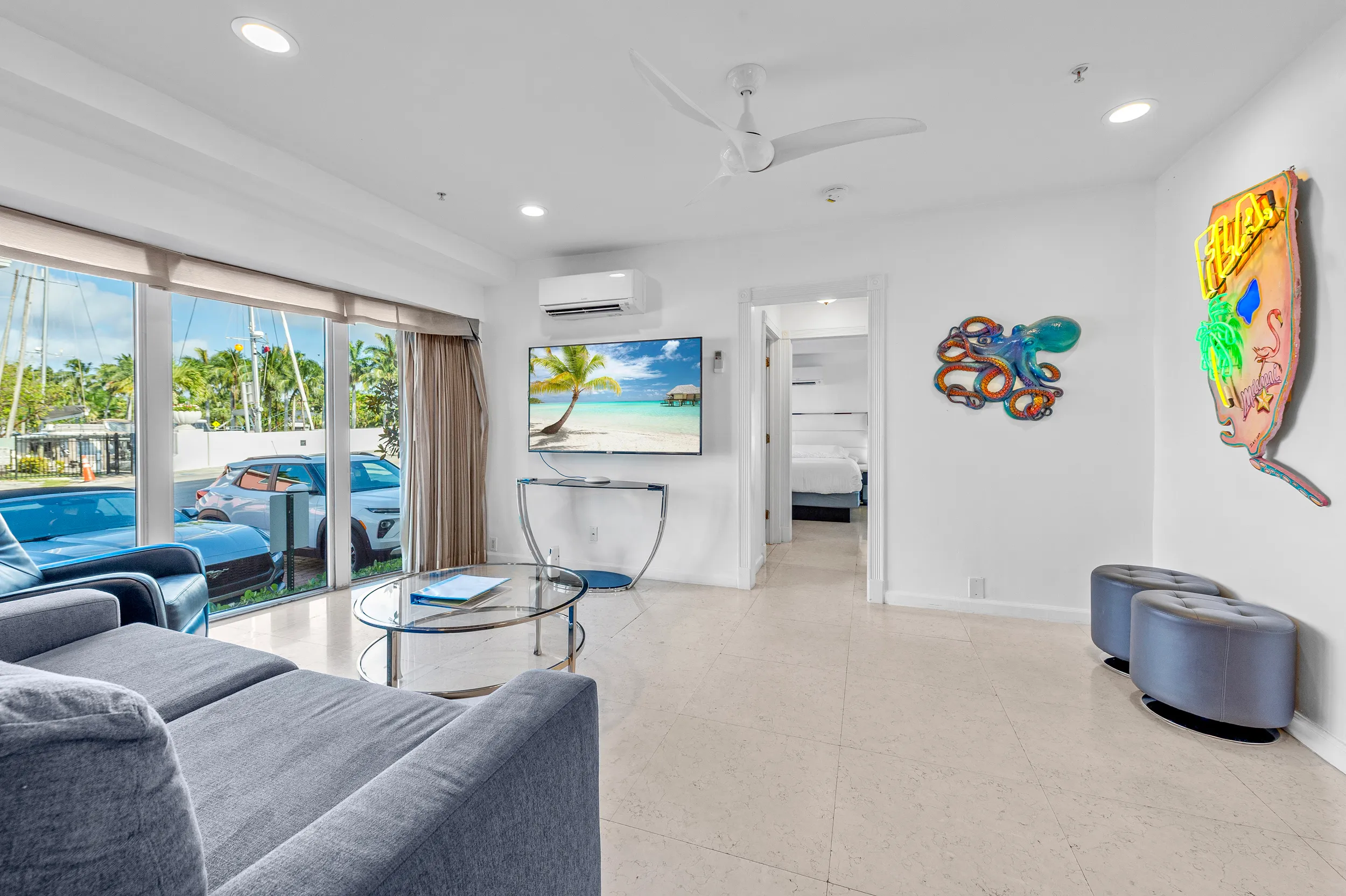 Modern living room with large window showing parked cars outside, gray couch, glass coffee table, TV on wall, and two round stools under colorful wall art.