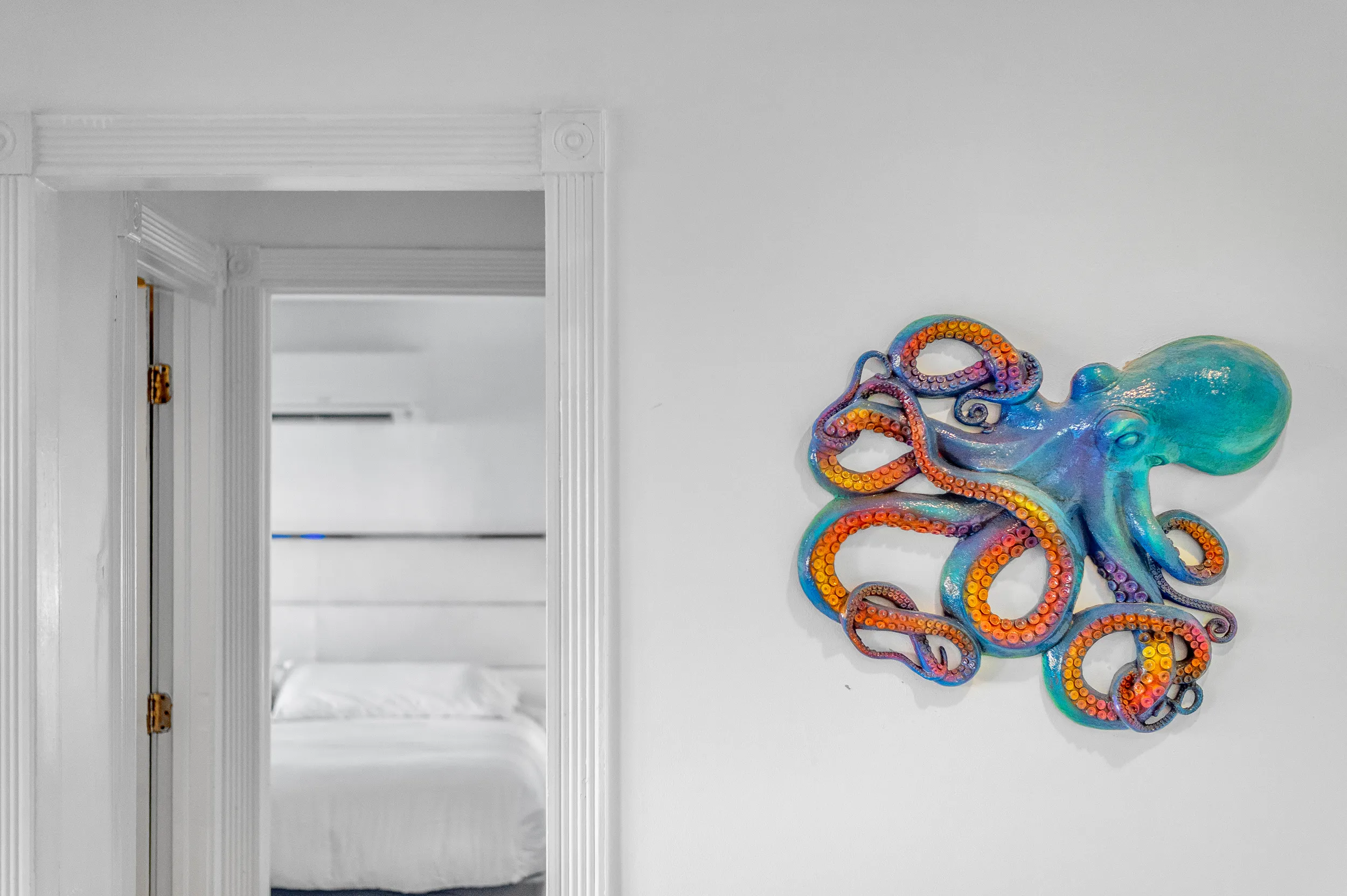 Colorful octopus wall sculpture mounted on white wall near doorway to bedroom with white bed.