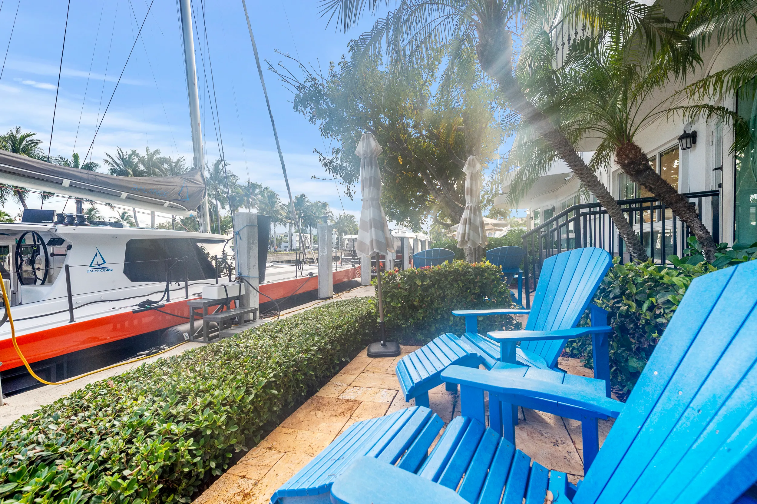 Blue Adirondack chairs and closed umbrellas on a patio by a marina with a moored sailboat and palm trees.