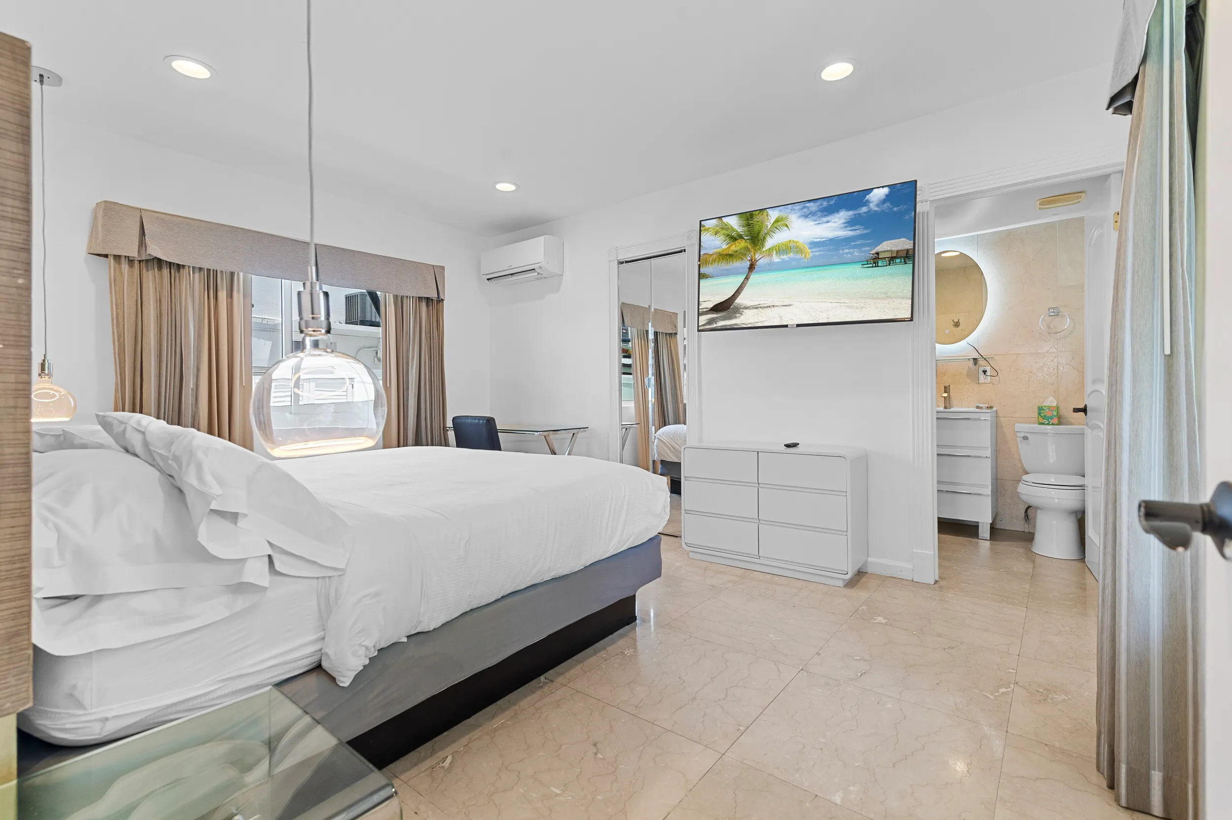Bright hotel room with a large bed, glass pendant light, TV displaying a beach scene, and an open door leading to a bathroom.