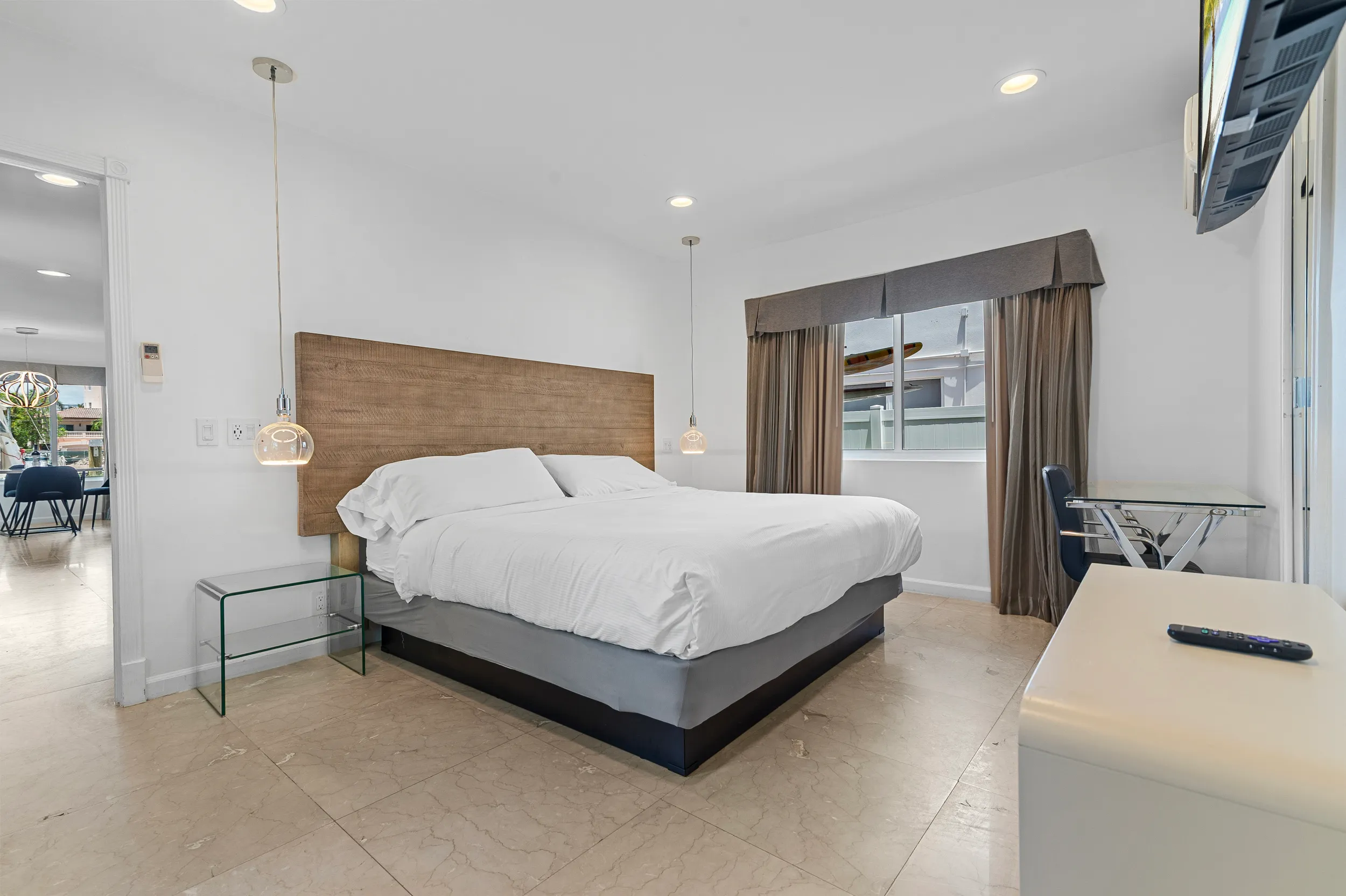 Minimalist bedroom with a large bed featuring a wooden headboard, white bedding, glass nightstand, and a window with brown curtains.