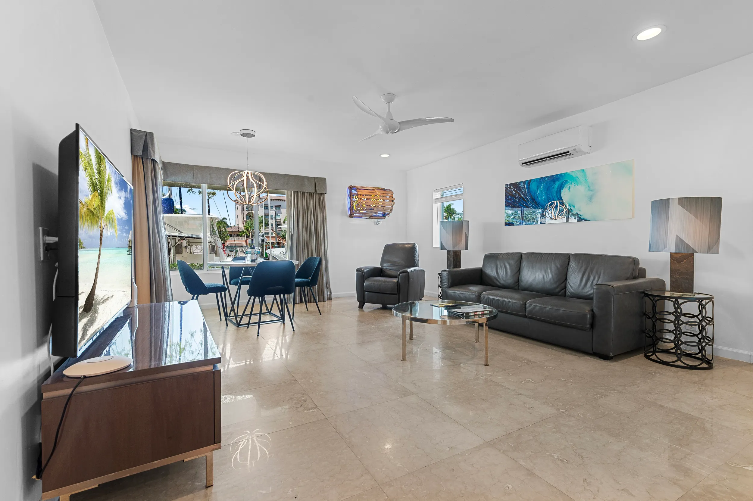 Modern living room with leather sofa, armchair, round glass coffee table, wall-mounted TV, dining table with blue chairs, and large windows showing outdoor greenery.