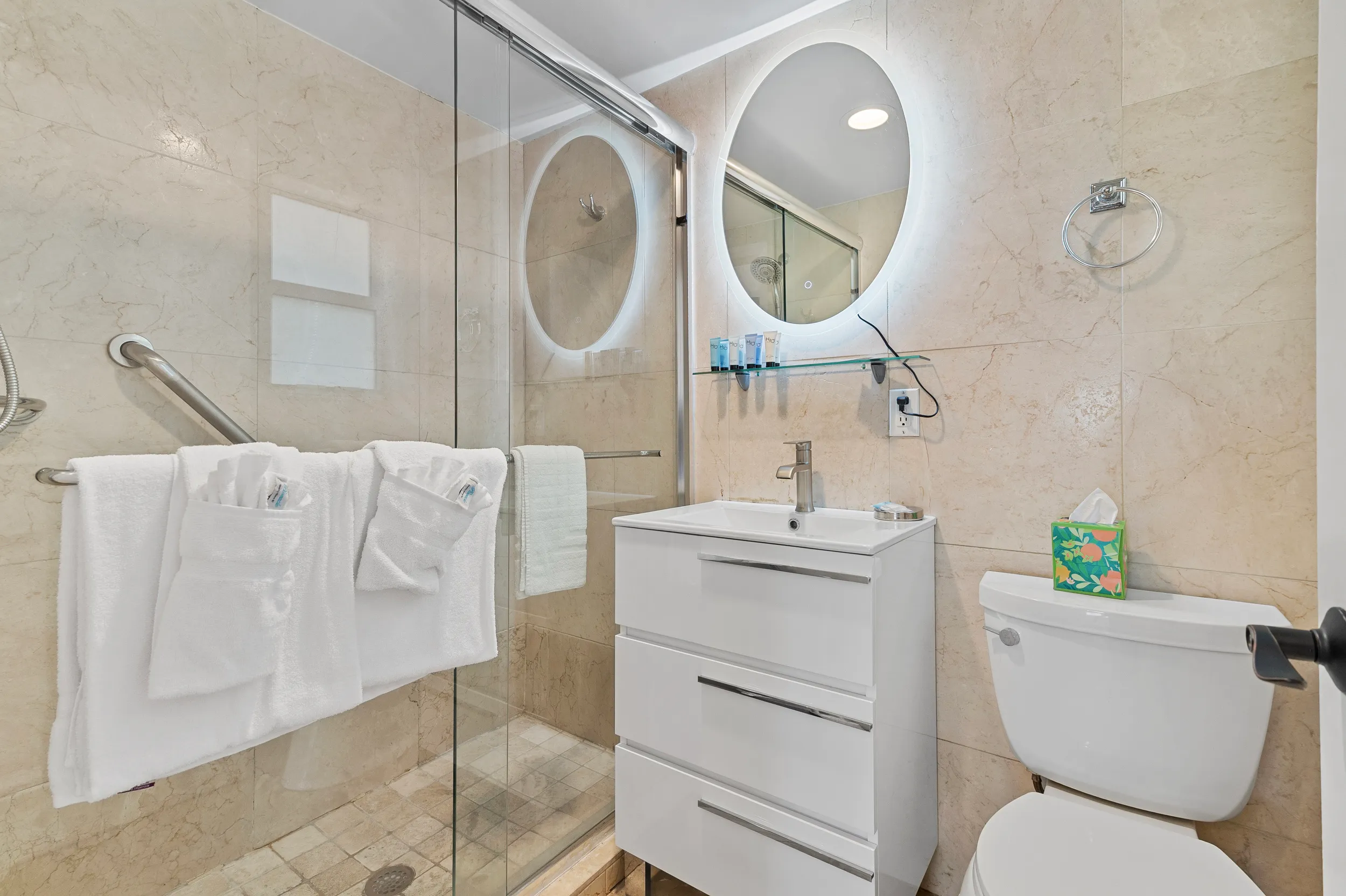 Modern bathroom with a glass shower, white vanity with illuminated oval mirror, and toilet with floral tissue box.