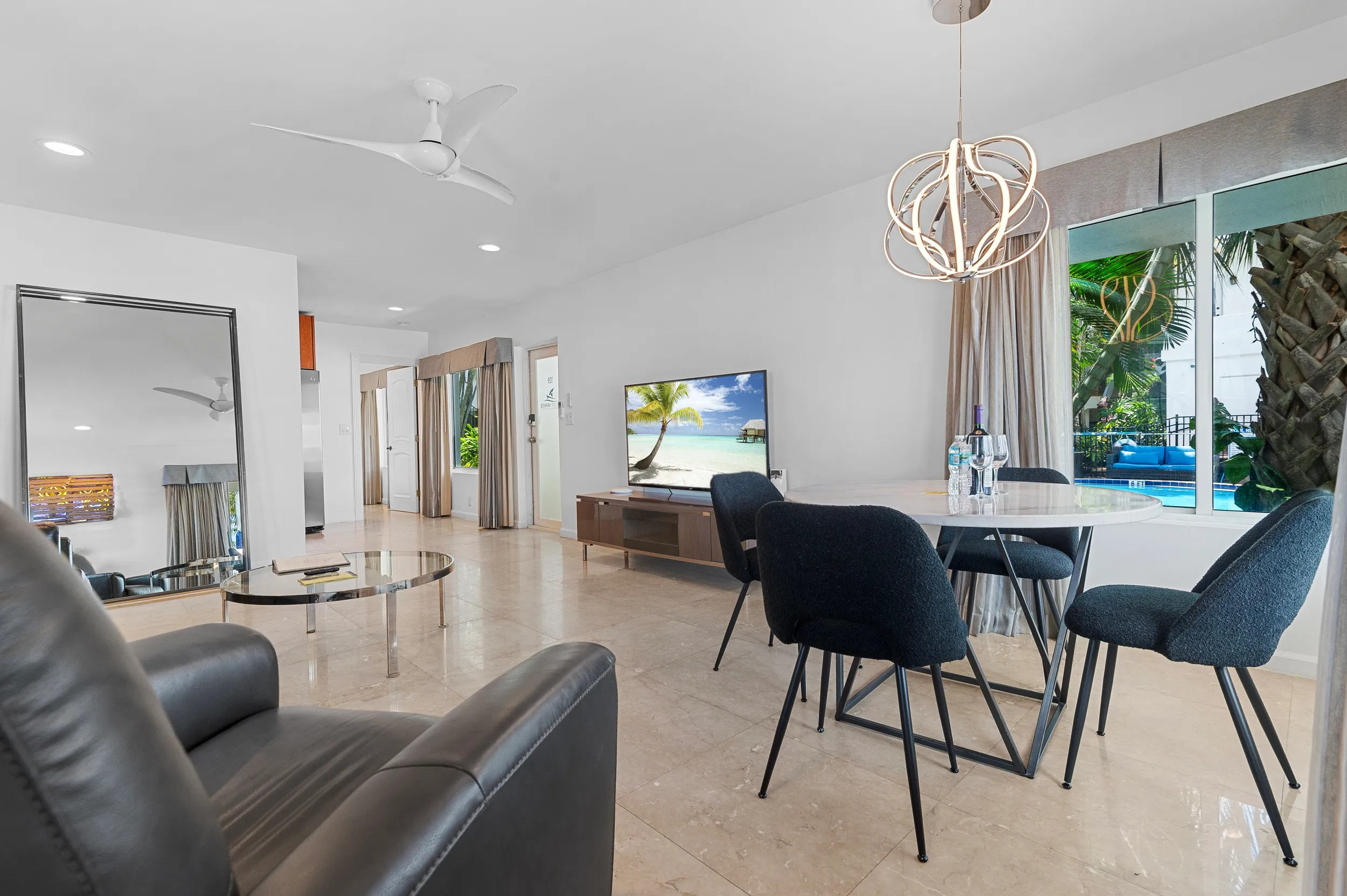 Modern living room with leather chair, glass coffee table, large mirror, round dining table with four black chairs, and TV showing a beach scene.
