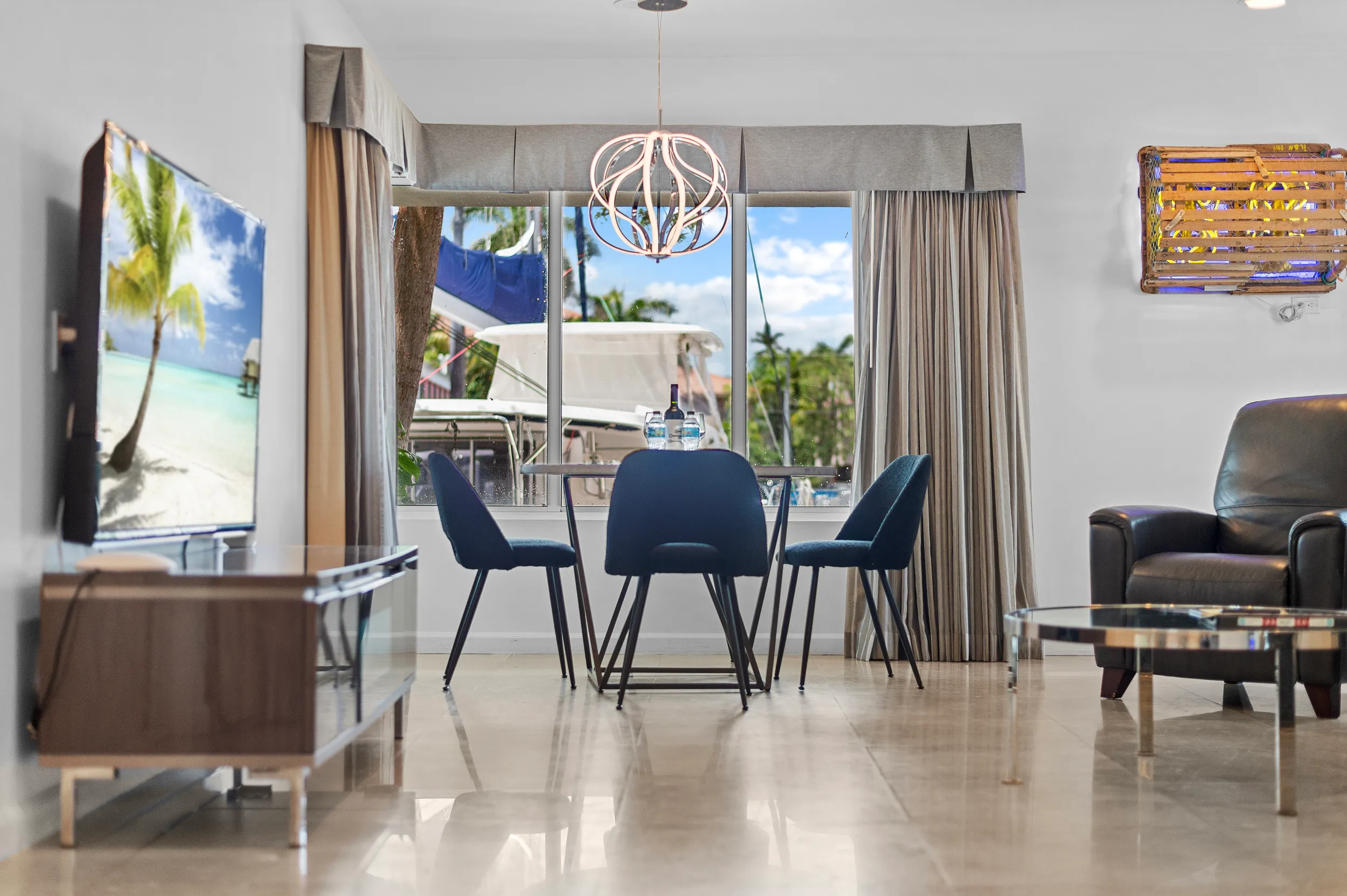 Modern dining area with a round table, four blue chairs, a bottle and two water glasses, a large window overlooking boats outside, and a living room with a black armchair and a round coffee table.