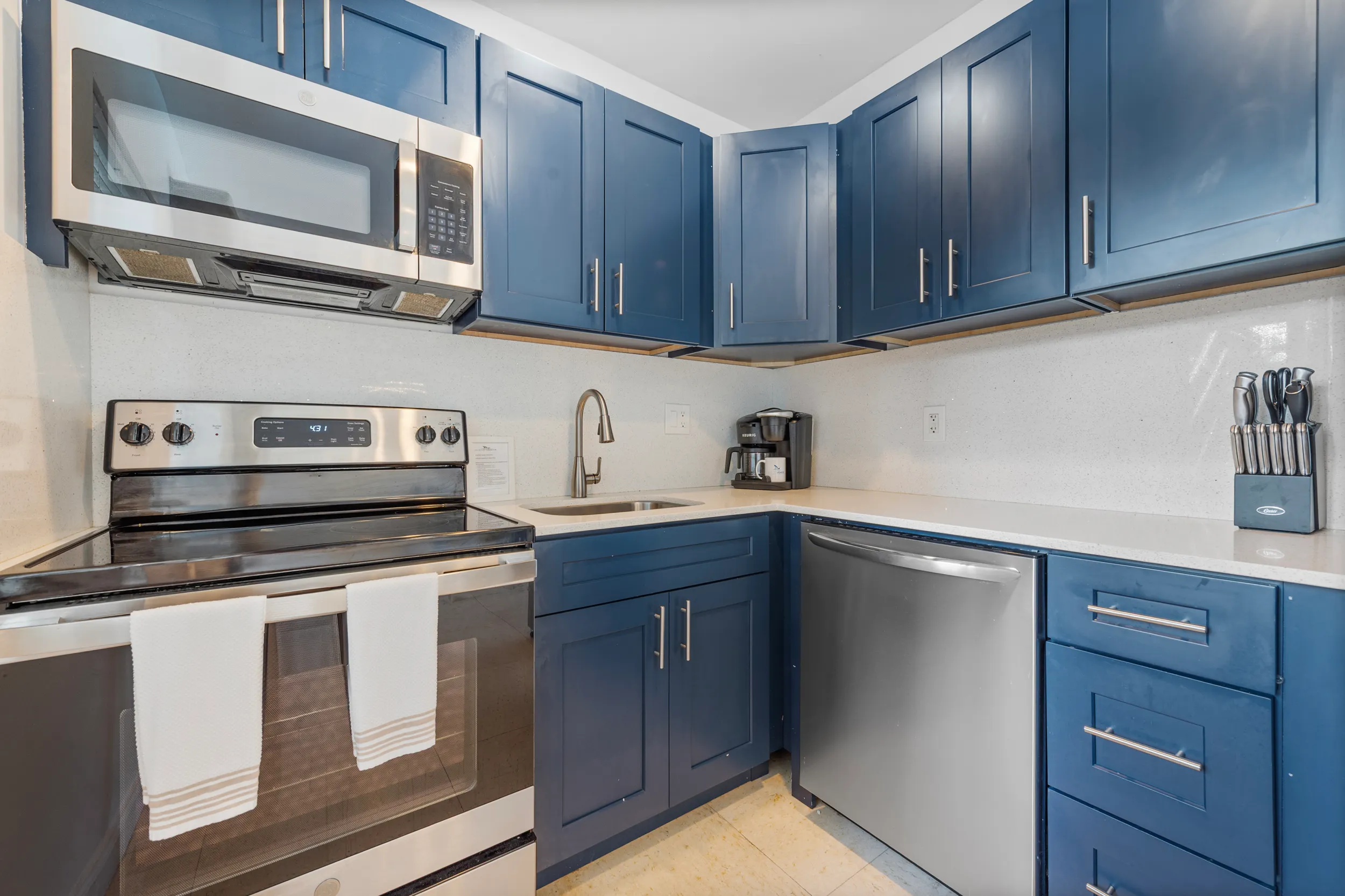Modern kitchen corner with blue cabinets, stainless steel oven, microwave, dishwasher, sink, coffee maker, and a knife block on white countertops.