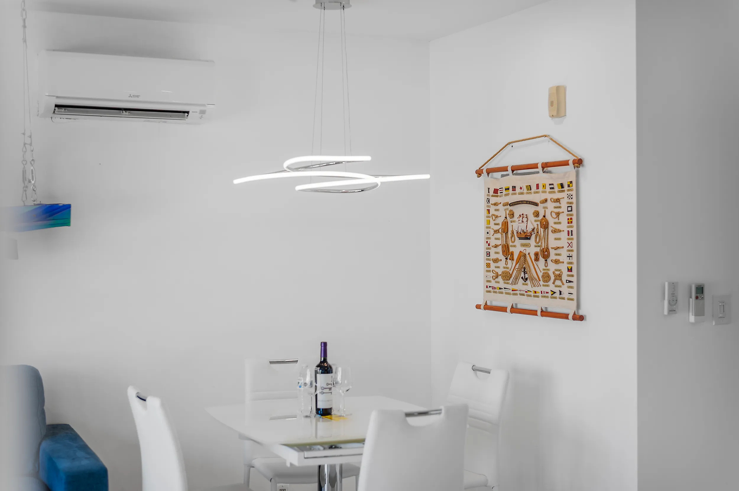 Minimalist dining area with a white table, four white chairs, a modern circular pendant light, a bottle of wine with glasses on the table, and a decorative wall hanging with nautical flags and knots.