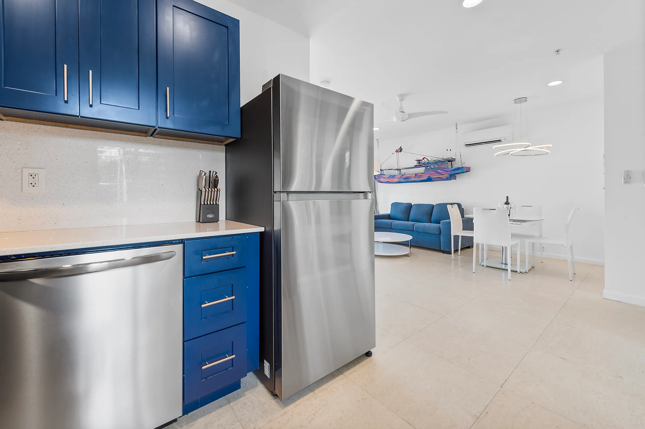 Modern kitchen with stainless steel refrigerator and dishwasher, blue cabinets, and an open living area featuring a blue sofa and white dining table with chairs.