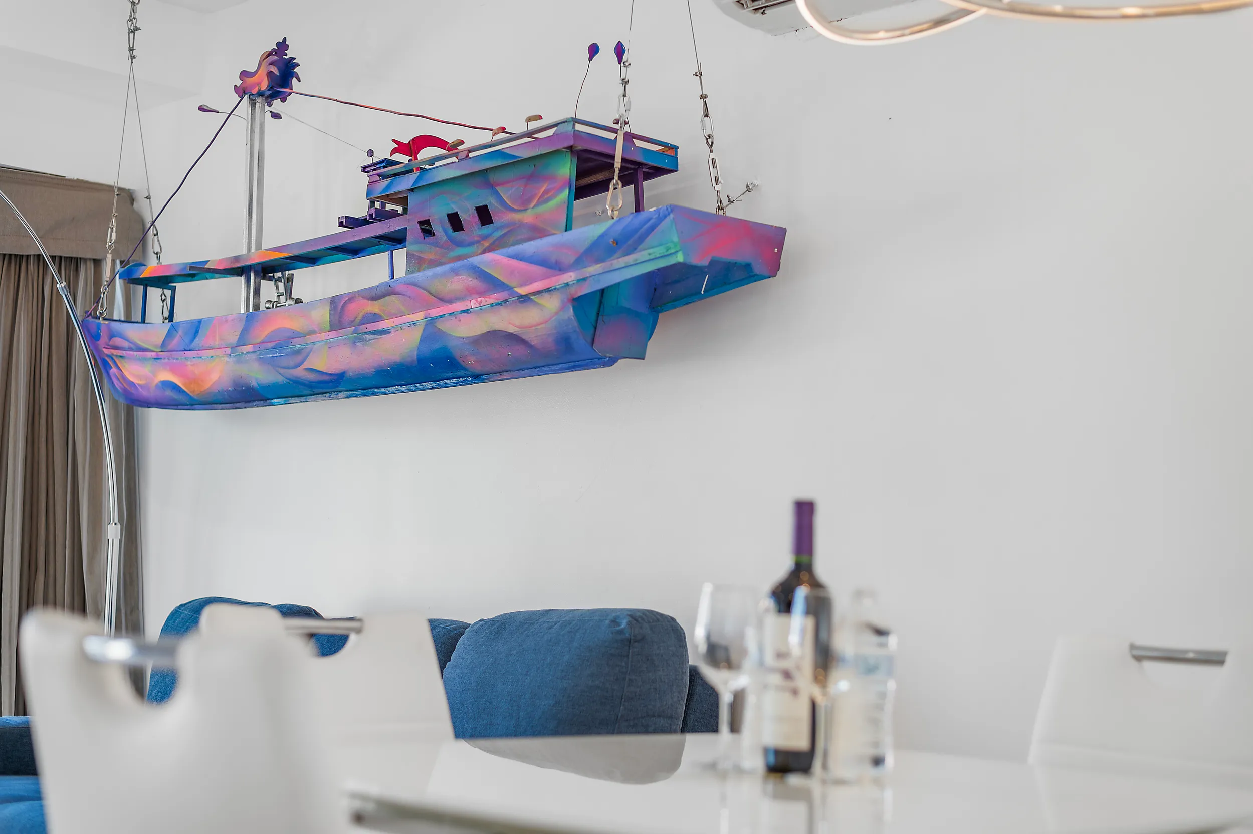 Colorful decorative boat model suspended from the ceiling in a modern living room with a dining table and a wine bottle with glasses.