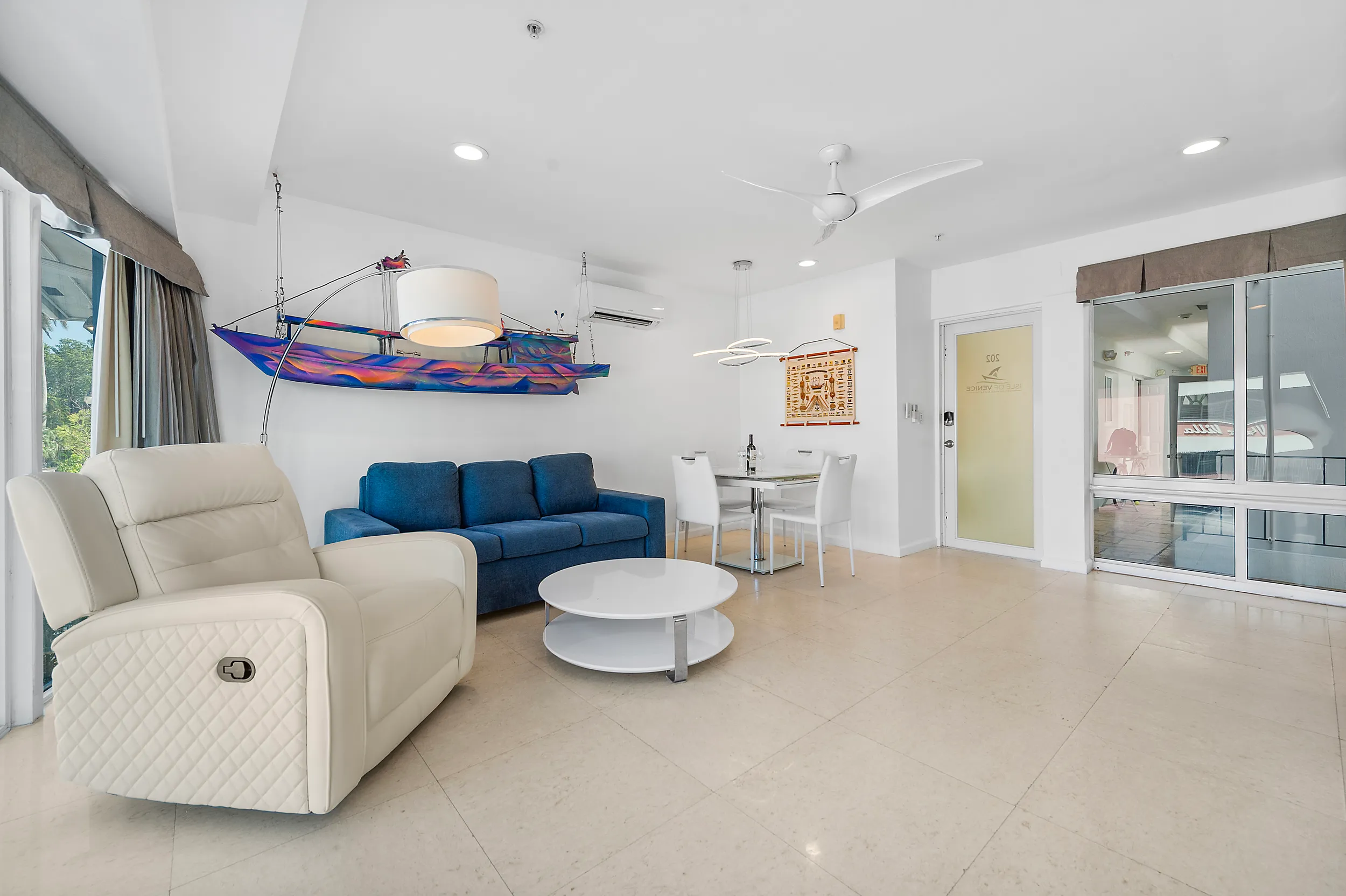 Modern living room with cream leather recliner, blue sofa, round white coffee table, and a small dining area with four white chairs under pendant lights.