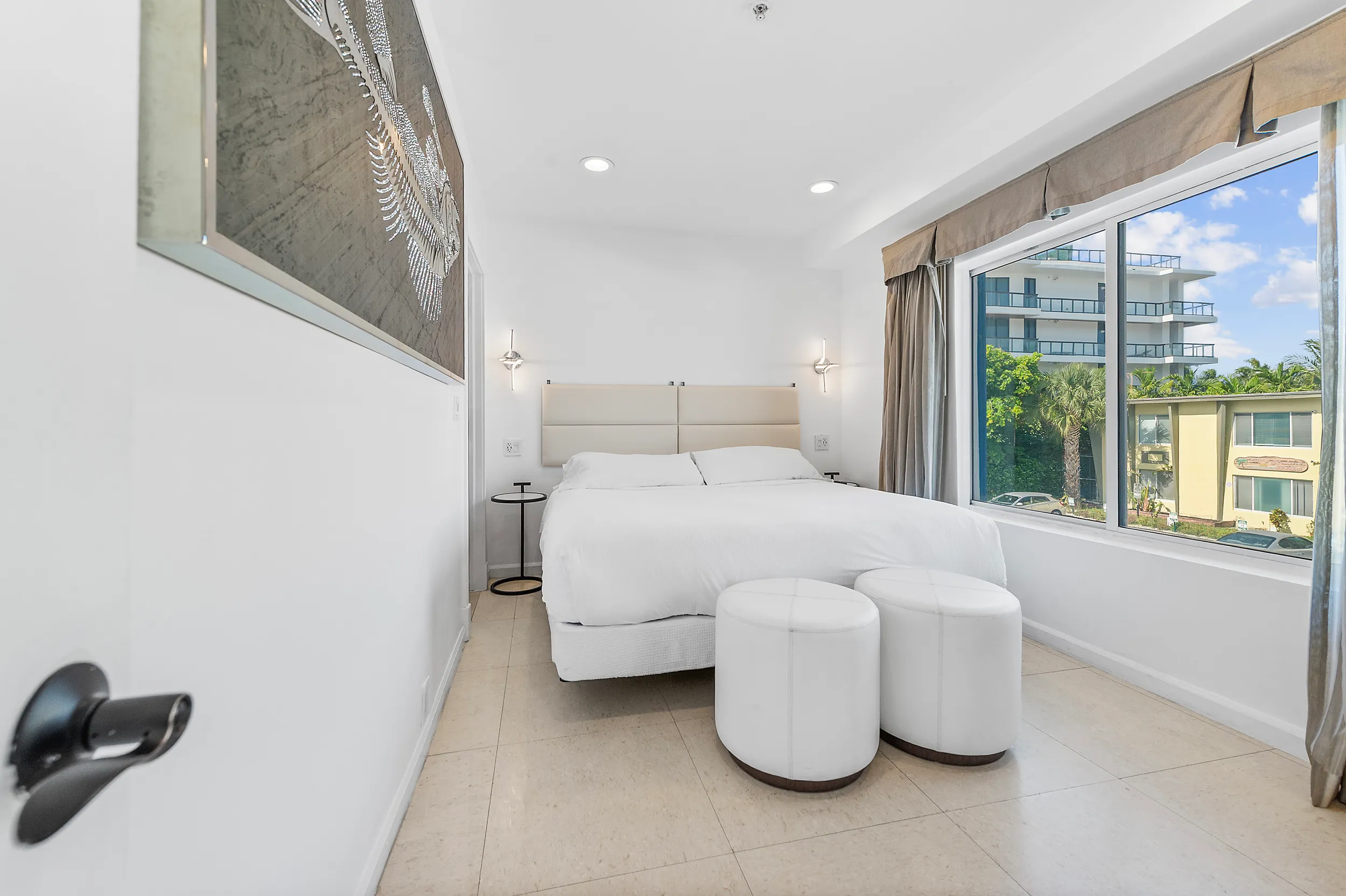 Bright bedroom with a large bed, two white ottomans, modern wall sconces, and a window showing buildings and palm trees outside.