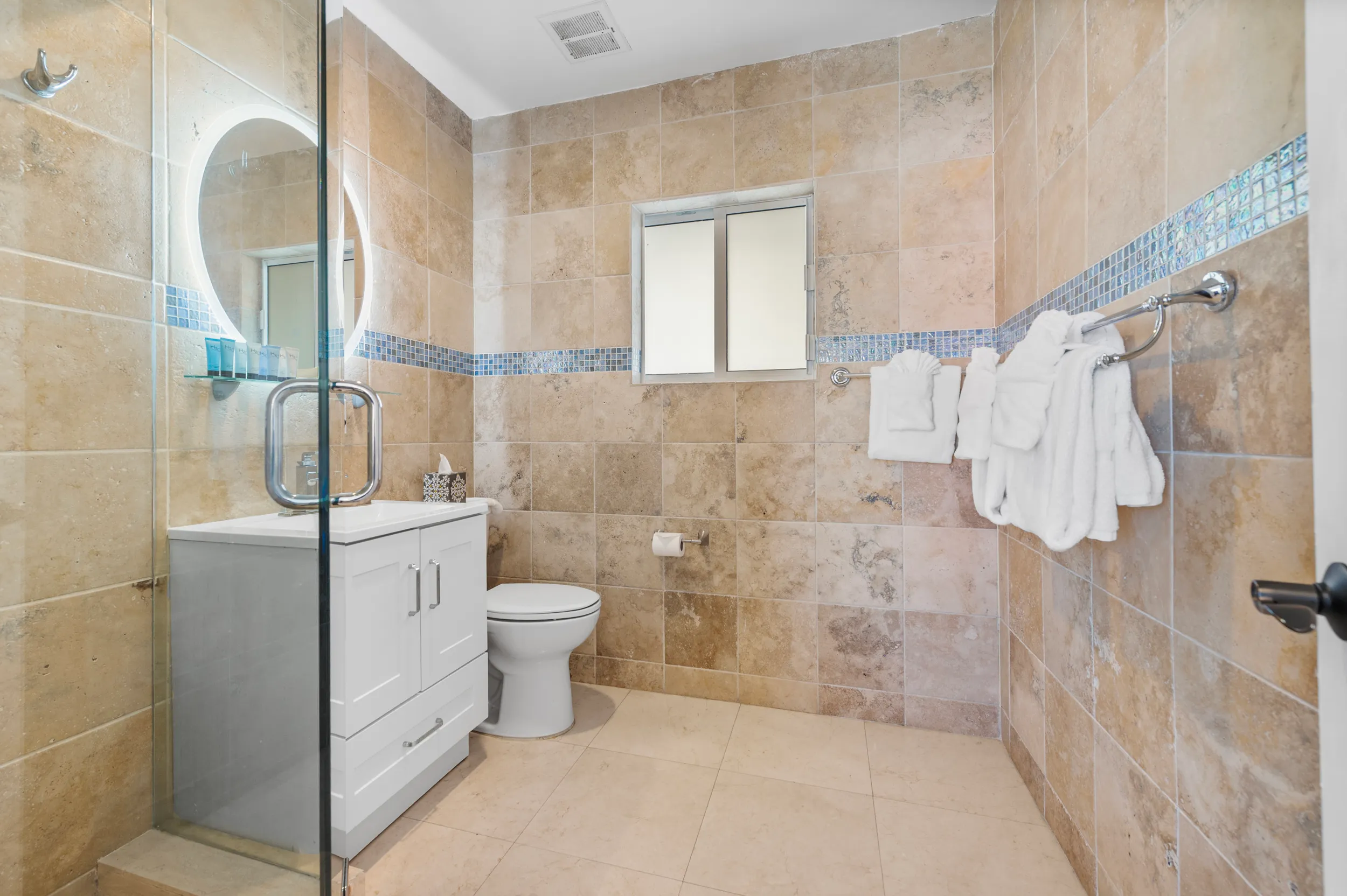 Bathroom with beige tiled walls and floor, white vanity with illuminated round mirror, toilet, glass shower door, and towels hanging on a wall rack.