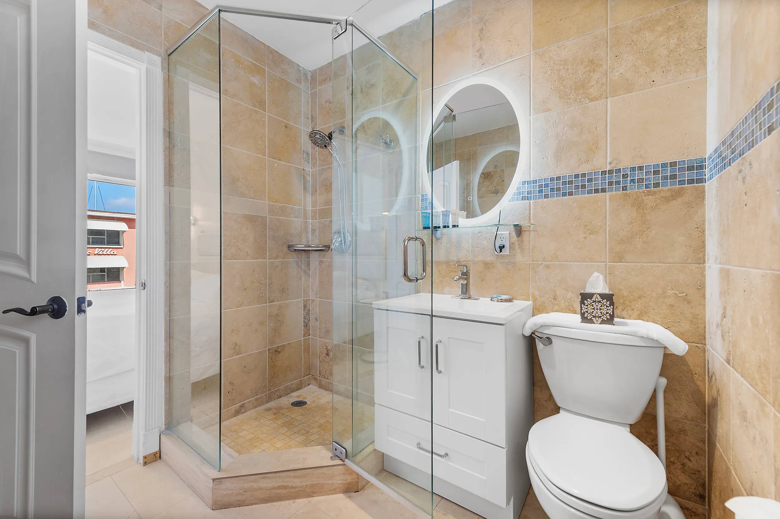 Small bathroom with tan tiles, glass shower enclosure, white vanity with round illuminated mirror, and toilet with tissue box on the tank.