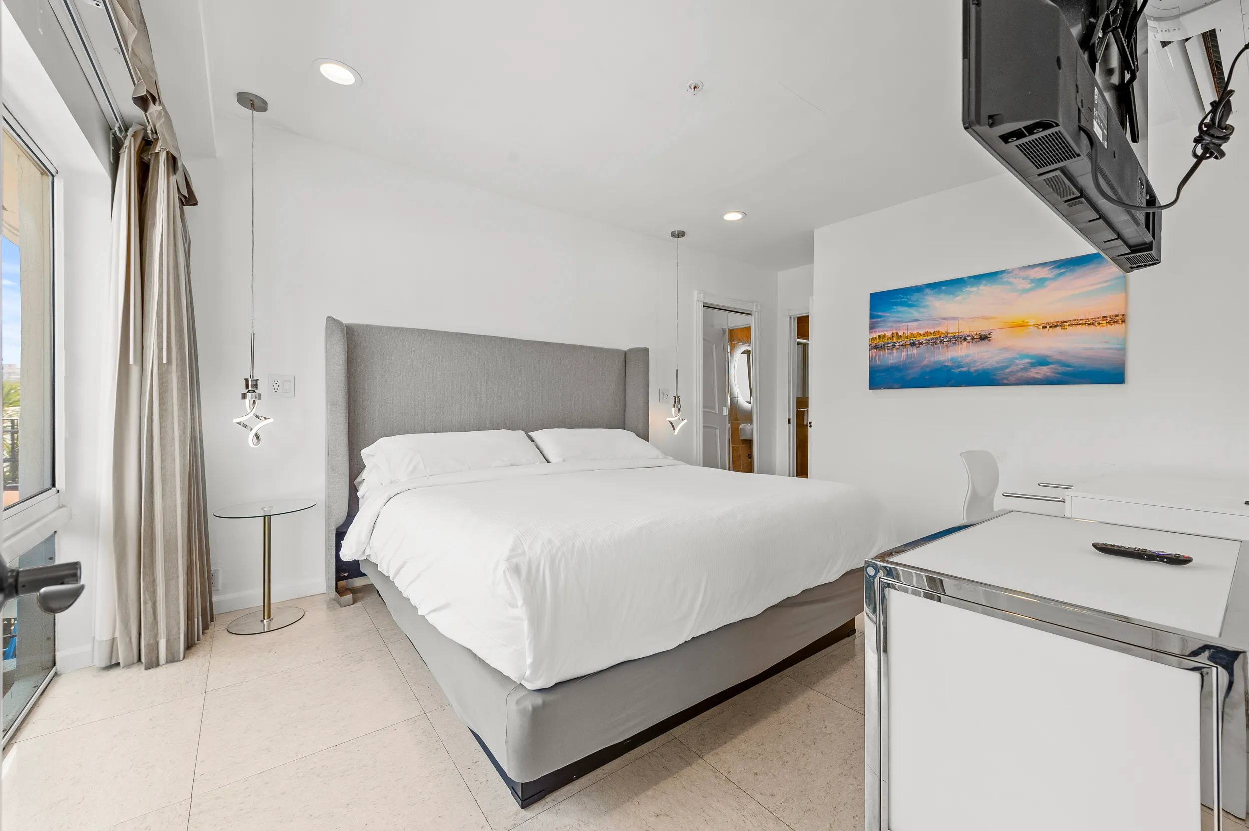 Modern bedroom with a large bed with gray headboard, white bedding, hanging pendant lights, glass side table, wall-mounted TV, and a colorful landscape painting.