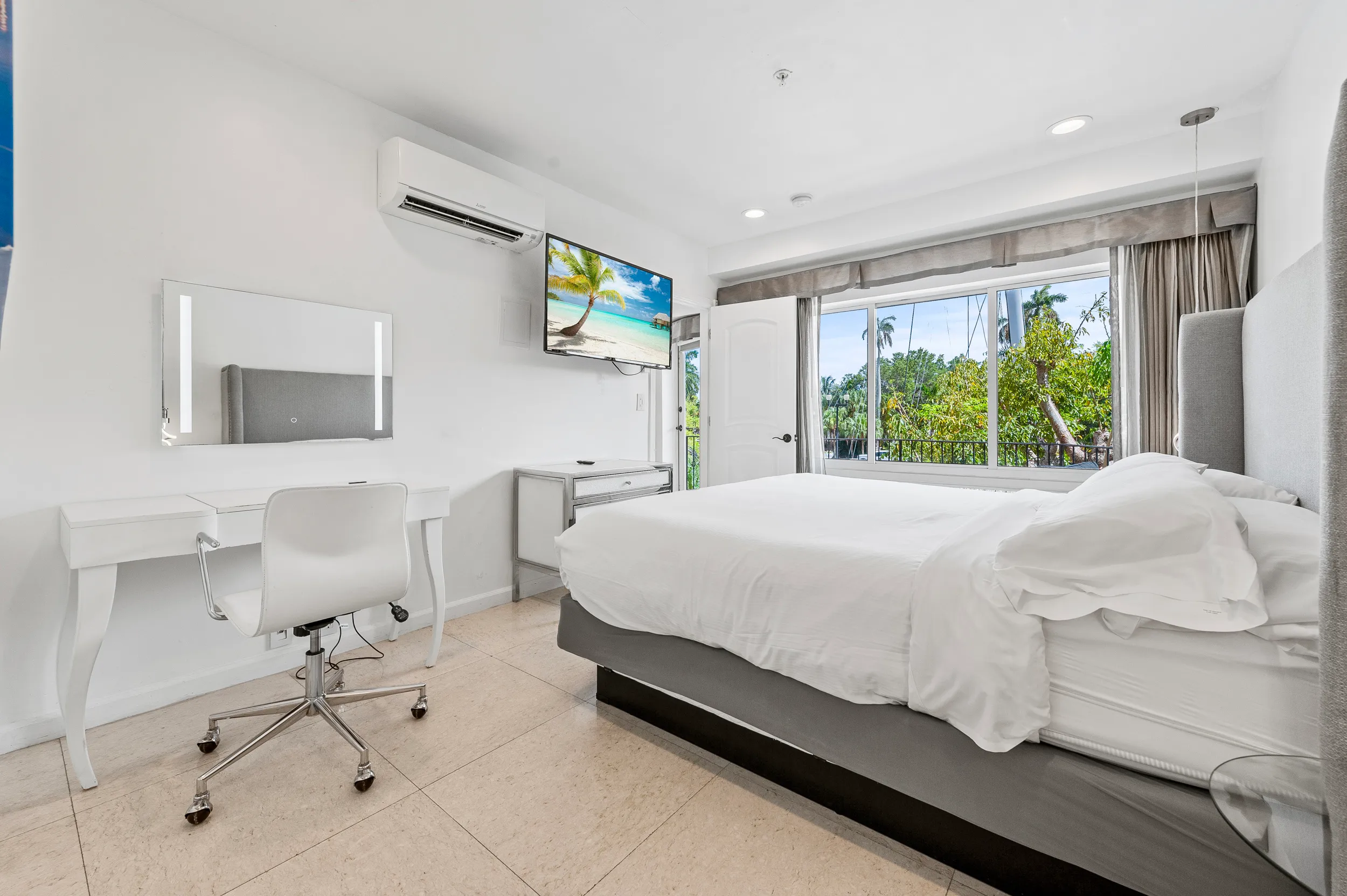 Bright, minimalistic bedroom with a white desk and chair, wall-mounted TV, large window overlooking greenery, and a bed with white linens.
