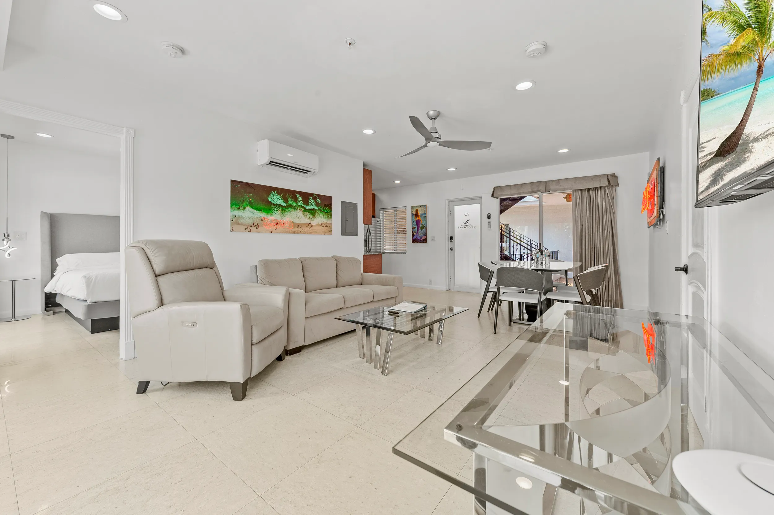 Modern living room with beige leather armchair and sofa, glass coffee table, ceiling fan, dining area, and wall-mounted TV showing a beach scene.