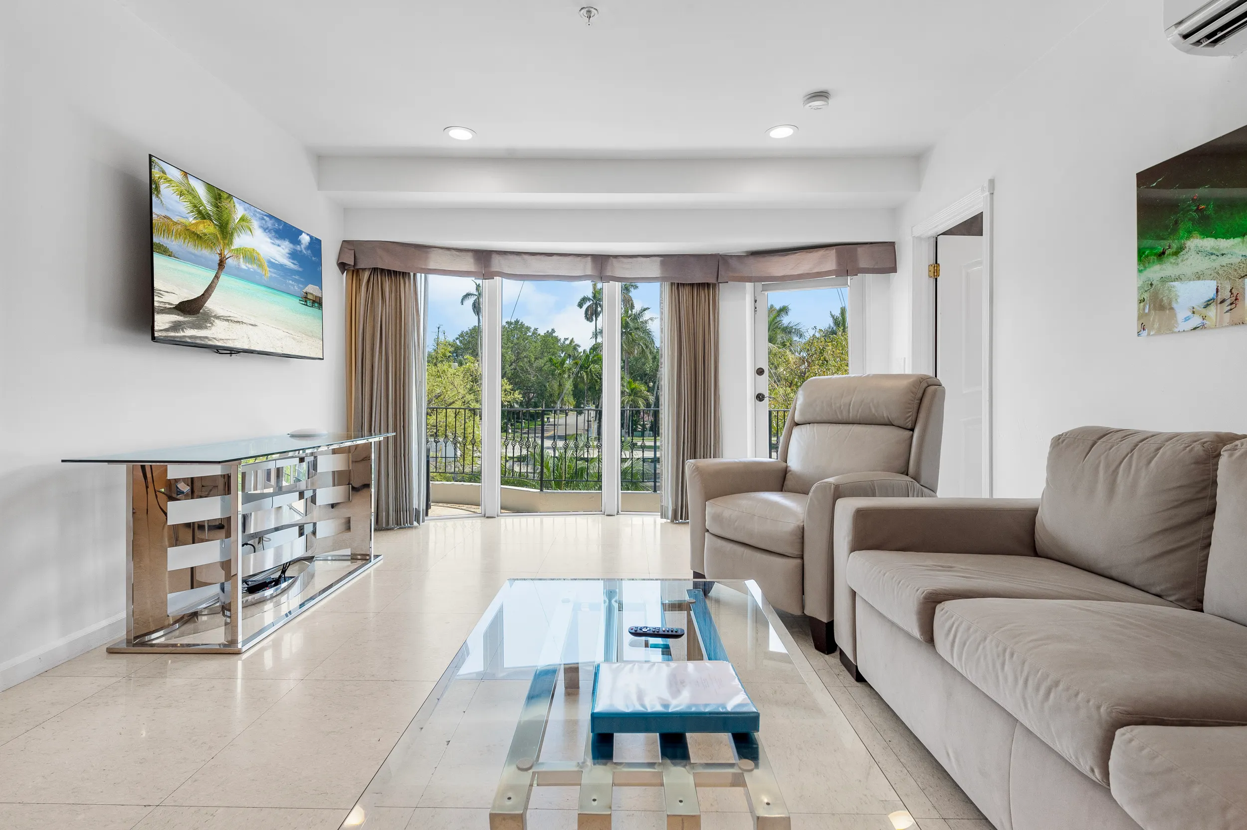 Modern living room with beige sofa and armchair, glass coffee table, flat-screen TV, and large windows showing green trees outside.